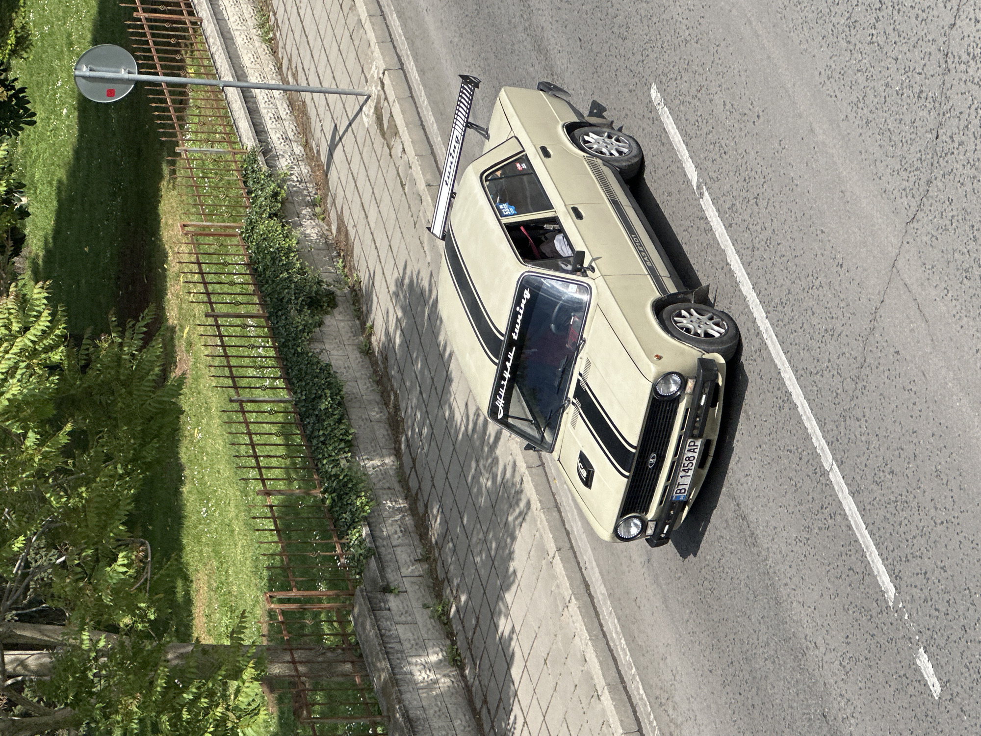 An elevated, high-angle shot shows a light yellow VAZ-2101 car with black racing stripes and a large, black and white racing spoiler parked on the side of a road in Veliko Tarnovo, Bulgaria. The car is situated on the edge of a paved area with gray concrete blocks, alongside a road with a solid white line marking its edge.
To the left of the paved area is a low fence made of dark brown metal bars, bordering a green area with grass and some trees. A round, gray traffic sign on a pole stands near the fence.
The car's license plate reads "BT 1458 AP." "Missile tuning" is written on the upper left part of the car's windshield. The driver's side door is open, revealing the red interior. The environment suggests a residential area.