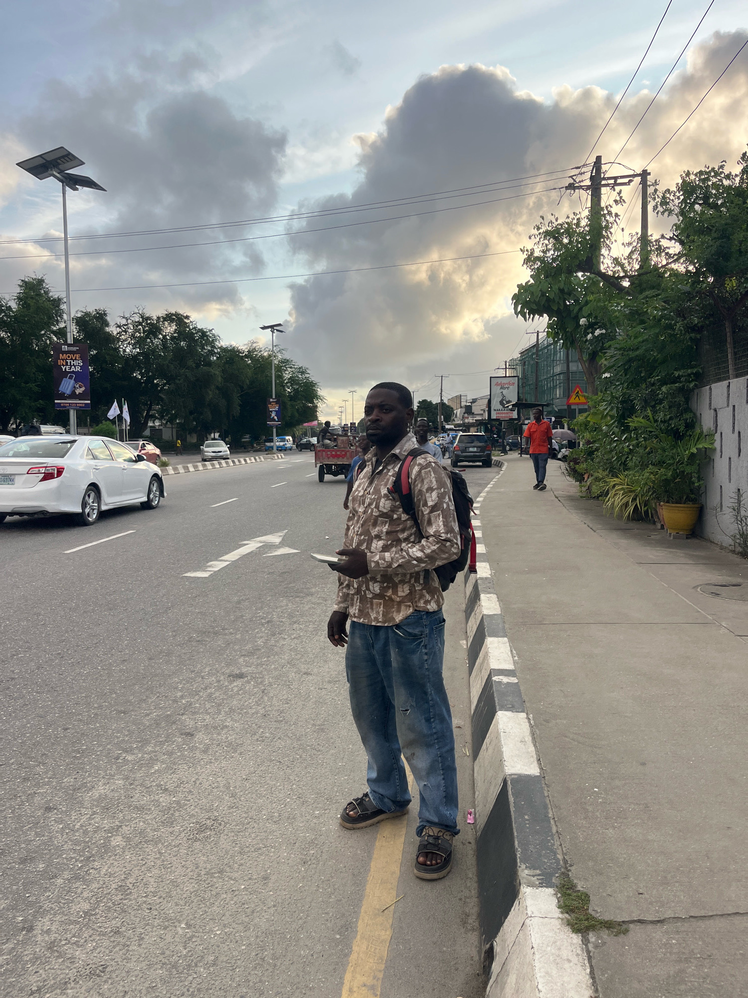 The image depicts a street scene in Lekki Phase I, Nigeria, under a cloudy sky. A man with dark skin, wearing a patterned long-sleeved shirt, distressed blue jeans, and dark sandals, stands on the sidewalk. He has a dark backpack with red straps. In his right hand, he holds a white object, possibly a phone. He is facing towards the camera, with a slight gaze to his left.
The sidewalk is bordered by a curb painted with black and white stripes. Beyond the curb is a multi-lane asphalt road with white lane markings and arrows. Several cars are visible on the road, including a white Toyota Camry on the left and other vehicles further in the distance. A red cart being pulled by an unseen source is also on the road, with a person standing in it.
On the right side of the sidewalk, there is a concrete wall with a rough, textured finish. Lush green plants in yellow and other planters are arranged along the wall, providing a contrast to the urban environment. Trees line the background on both sides of the road, with some bearing green foliage. A prominent billboard with an advertisement is visible on the left side of the road, and a traffic sign with a yellow warning triangle is present further down the street on the right. Electrical wires and poles are also part of the overhead infrastructure. The overall lighting suggests it is either early morning or late afternoon.