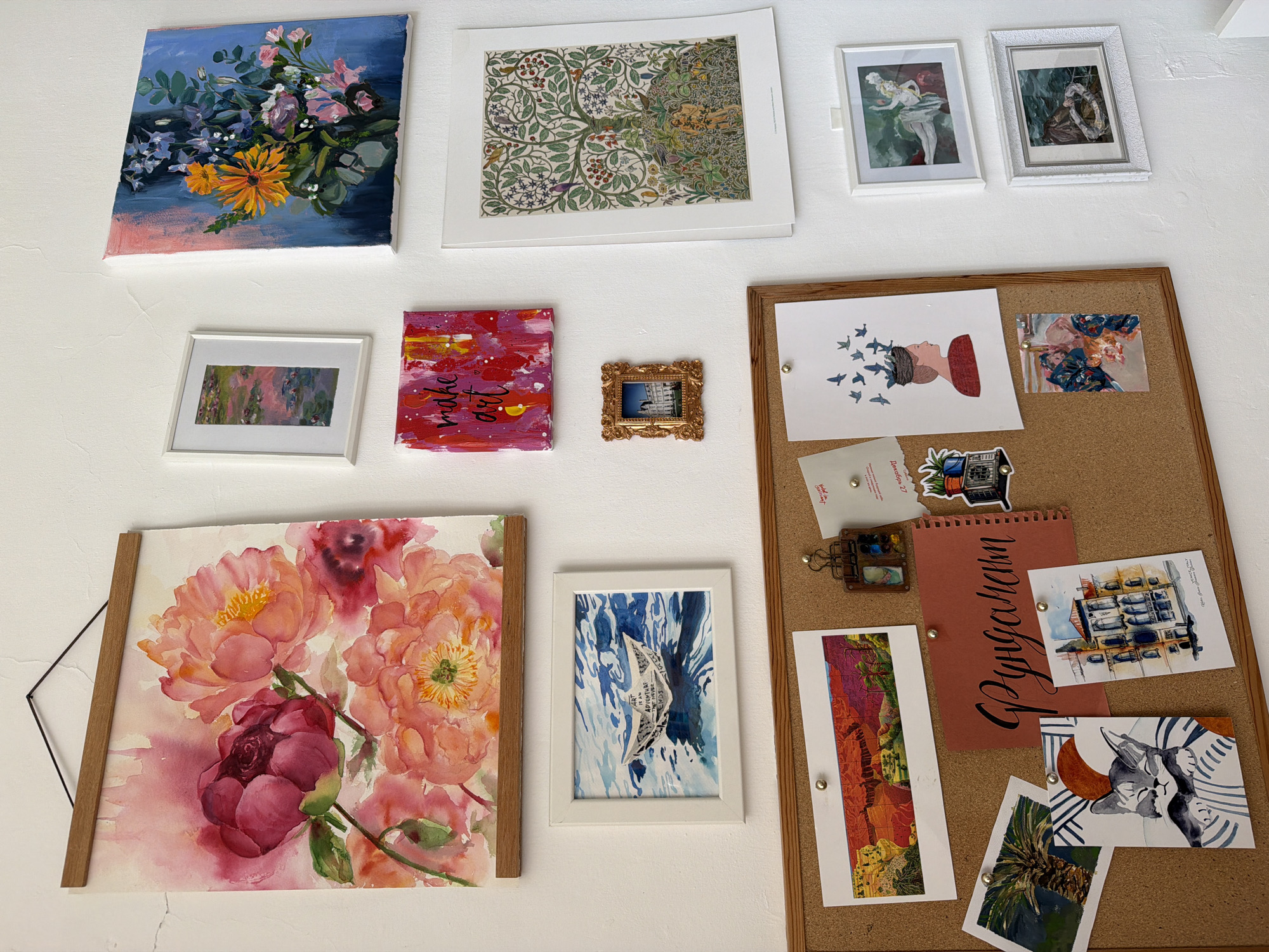 A collection of artwork and decorative items are arranged on a white wall. The collection includes a canvas painting of flowers, framed floral design print, two small framed art prints, a framed painting of water lilies, an abstract painting with the words "make art", a small ornate framed picture, a framed floral watercolor, and a framed painting of a paper boat on water. A corkboard is also mounted on the wall, displaying various items, including postcards, drawings, a small notebook, stickers, and other paper ephemera. The location appears to be indoors, in a private home or art studio. There is no information to confirm this image is in Bat Yam, Israel.