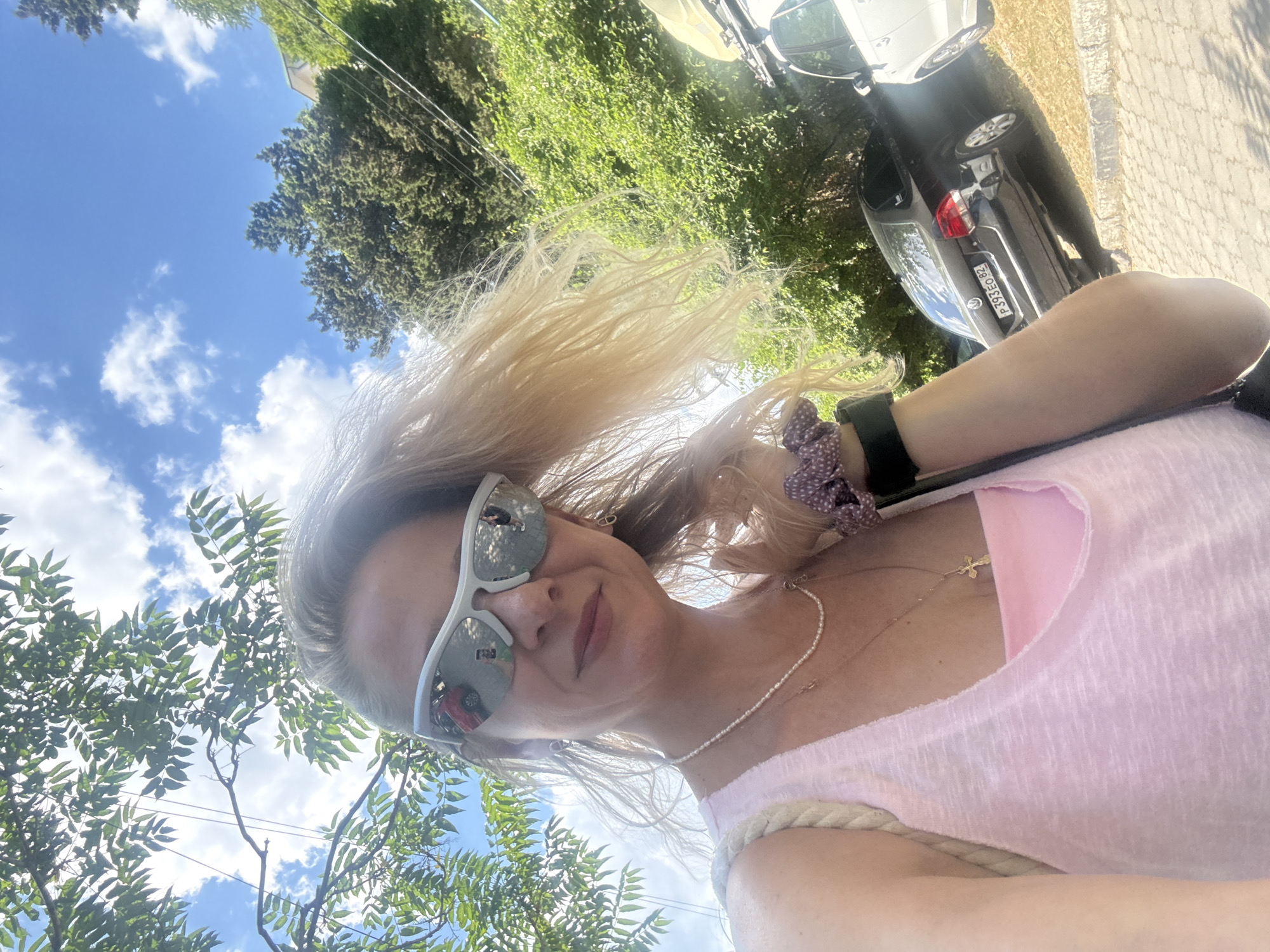 A woman with blonde hair and sunglasses takes a selfie outdoors, holding her hair with her right hand. She is wearing a pink tank top with a pink bra underneath, a pearl necklace with a cross pendant, and hoop earrings. Her sunglasses reflect a red car and a person. The background shows trees, a blue sky with clouds, and parked cars, including a black Volkswagen with a license plate starting with P393EO. The ground is paved with interlocking stones.
