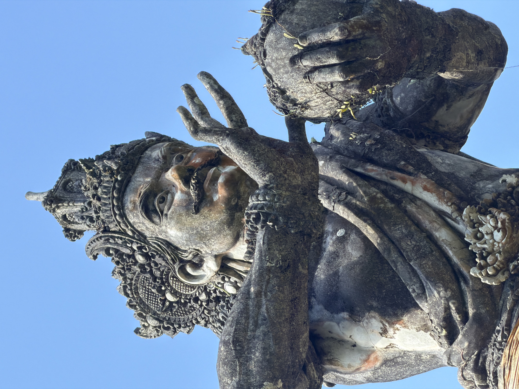 A close-up, low-angle shot shows the weathered, stone statue of a deity against a clear blue sky. The statue, likely located in Tampaksiring, Indonesia, depicts a figure with a stern expression, a prominent mustache, and an ornate headdress. Its body is adorned with draped clothing and bracelets. In its hands, the statue cradles a spherical object, possibly a vessel or fruit, with small plants growing on its surface. The stone exhibits signs of age and exposure to the elements, with patches of discoloration and vegetation.