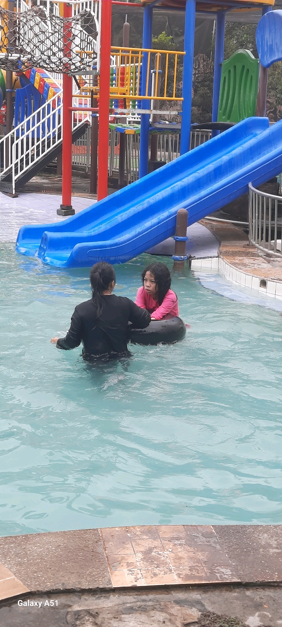 In Popoh, Indonesia, two individuals are present in a swimming pool. One is an adult wearing a black long-sleeved shirt, facing away from the camera and holding a black inner tube. The other is a child wearing a pink long-sleeved shirt, sitting in the inner tube and facing the adult. The pool water is clear and blue. Behind them, a vibrant blue water slide curves into the pool, part of a playground structure with red and blue support beams, yellow railings, and climbing nets. The surrounding environment includes trees and foliage.