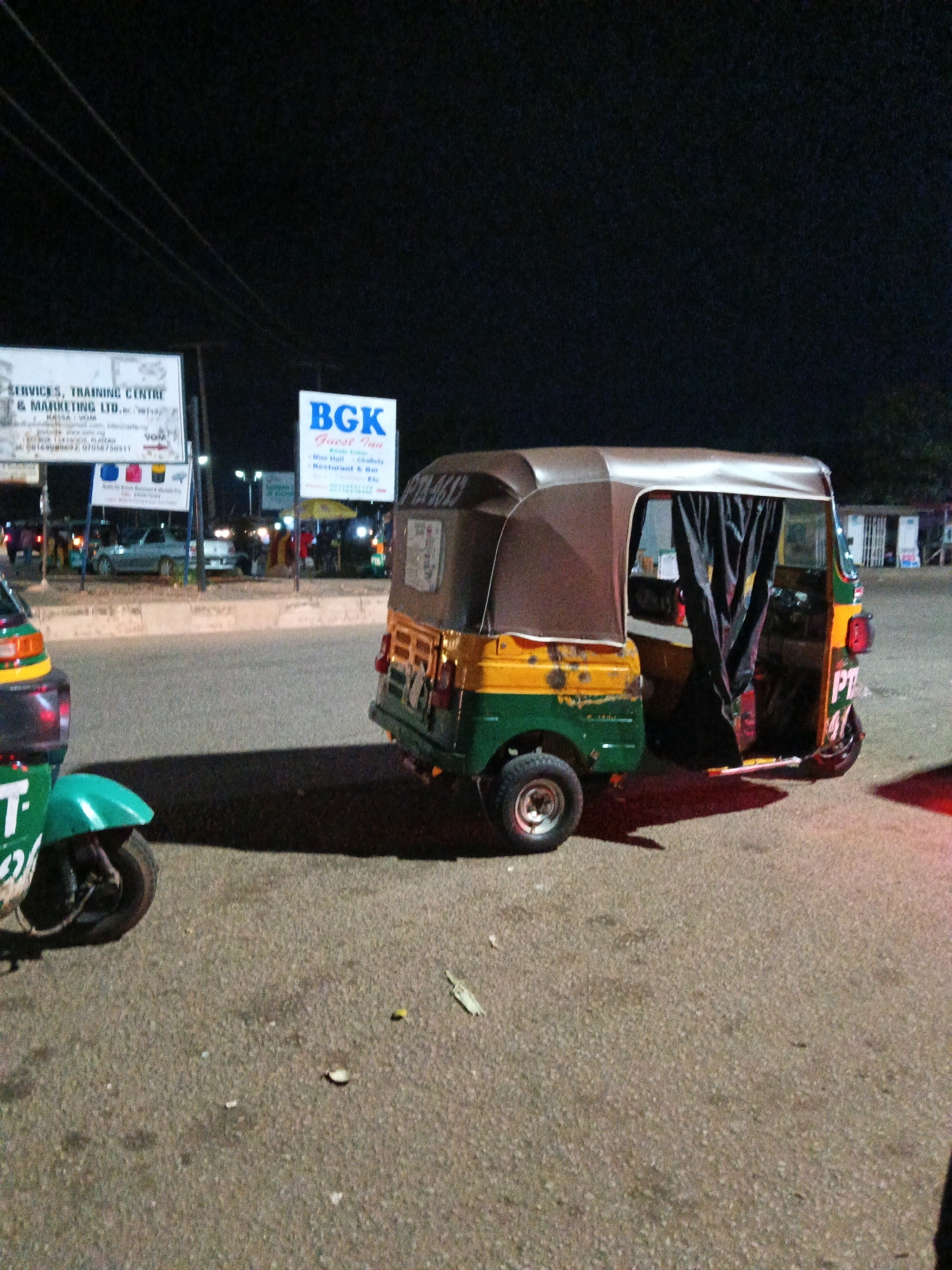 The image depicts a nighttime scene in Bukuru, Nigeria, likely near a marketplace or commercial area. In the foreground, a green and white auto-rickshaw is partially visible, facing left. Behind it and to the right, a yellow and green auto-rickshaw is parked, with its side curtain drawn. The auto-rickshaw has "PT 412" painted on its rear.
Further in the background, a white sign reads "SERVICES, TRAINING CENTRE & MARKETING LTD." Below this, another sign displays "BGK Forest Inn," with text indicating "Mini Hall, Chalets, Restaurant & Bar." Several other illuminated signs and structures suggest commercial activity. People are sparsely visible in the background, some appearing to be vendors or patrons. The ground is paved, and the lighting suggests artificial illumination from streetlights or commercial establishments. The overall environment appears to be an urban or semi-urban area during the evening.