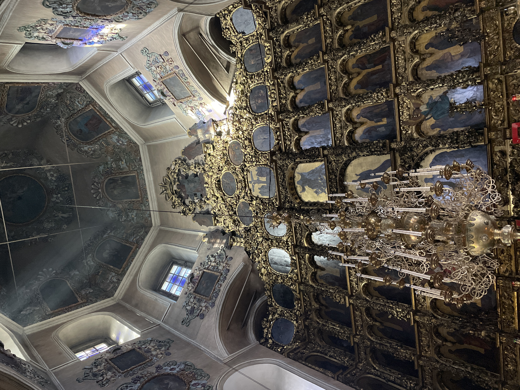 The image captures a highly ornate interior of what appears to be a church or cathedral, viewed from a low angle looking upwards. The ceiling features intricate painted decorations with floral and foliate motifs, surrounding smaller framed artworks or frescoes depicting figures. Several windows, some with stained glass in shades of blue, red, and yellow, are visible on the upper walls, allowing beams of natural light to stream into the space.
A dominant feature is a large, vertical wall or screen, reminiscent of an iconostasis, covered in multiple rows and columns of framed religious paintings or icons. These frames are heavily gilded and intricately carved, creating a dense, golden surface. The paintings within these frames primarily feature dark backgrounds with figures, many in a style suggestive of Eastern Orthodox iconography.
Two elaborate chandeliers are prominent in the foreground and mid-ground. One, closer to the viewer, is made of gleaming gold-colored metal with numerous arms designed to hold candles or lights, featuring delicate scrollwork. Another, partially visible, appears to have more silver-toned elements and transparent candle holders.
The overall impression is one of immense grandeur and detailed craftsmanship, with a rich palette of gold, dark blues/greens from the icons, and warmer tones on the ceiling and upper walls. Sunlight filtering through the windows highlights the dust in the air and adds a sacred atmosphere. There is no discernible activity or visible text in the image.