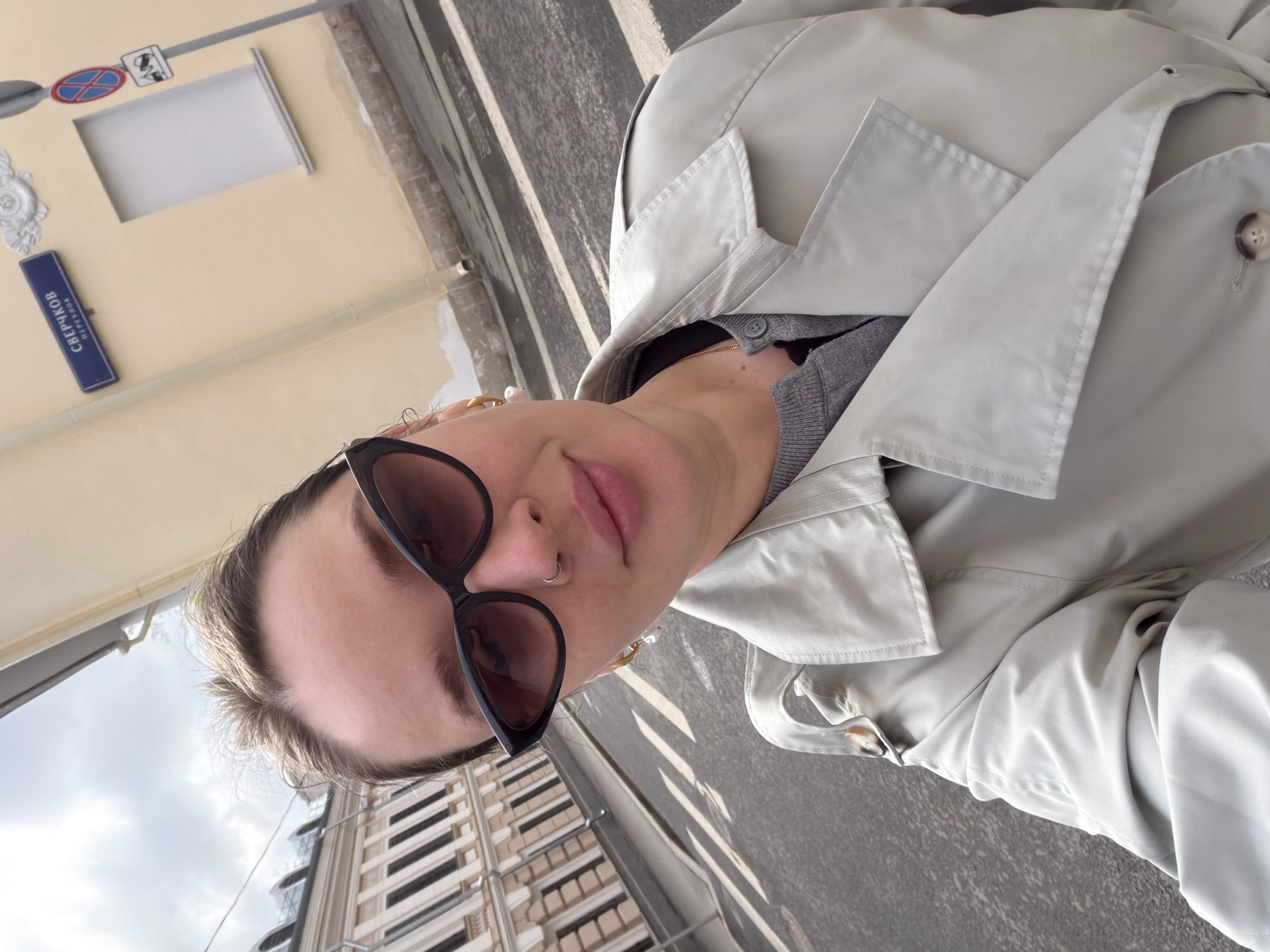 The image is a selfie of a person wearing sunglasses and a light-colored trench coat, likely in Moscow, Russia. In the background, there is a building facade, and street signage with Cyrillic script indicating the "Сверчков переулок" (Sverchkov Pereulok), which translates to Sverchkov Lane. A street sign is also visible. The photograph appears to have been taken outdoors during the day.