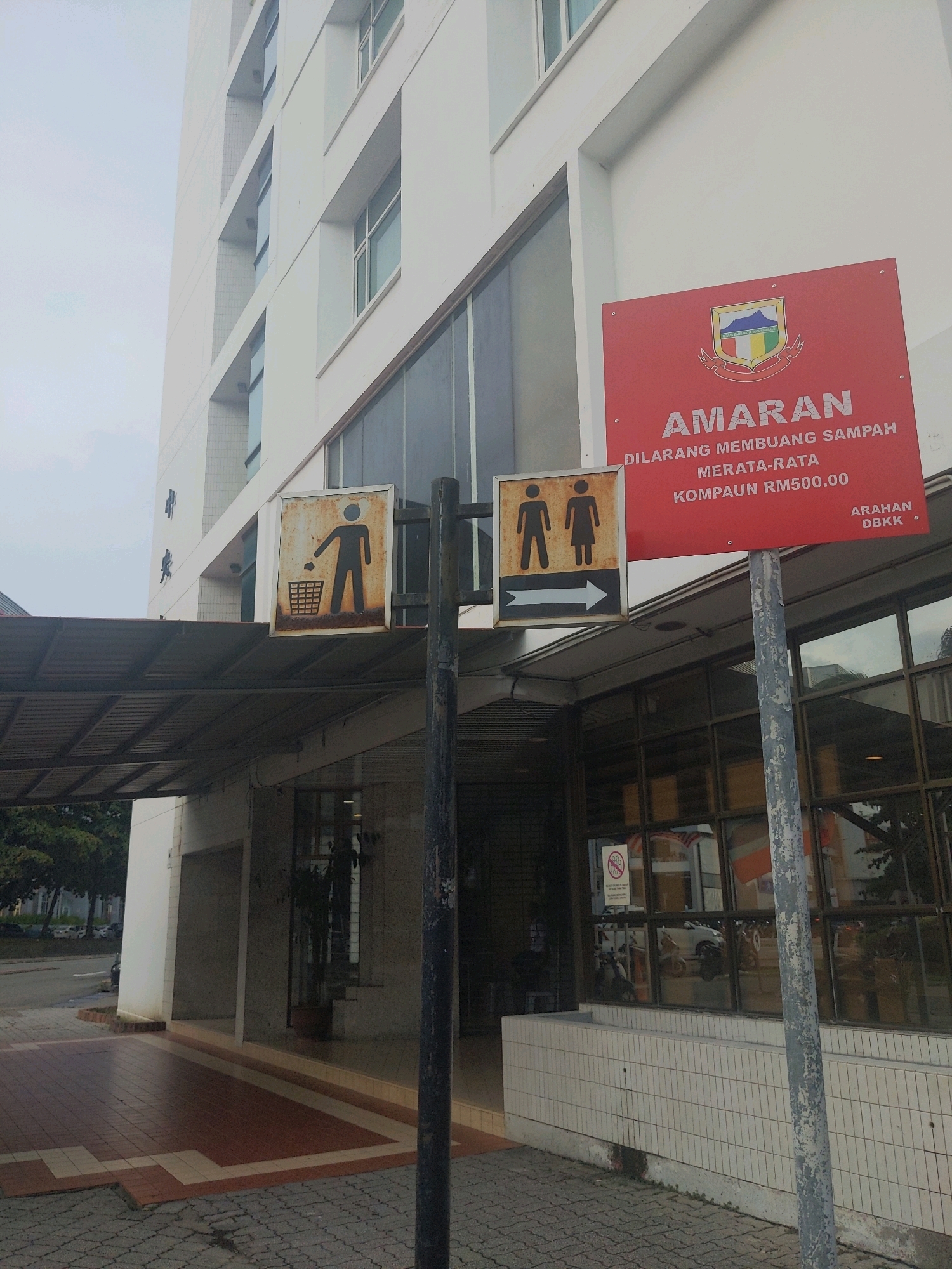 The image shows an outdoor scene in Kota Kinabalu, Malaysia, with a white building in the background and a paved walkway in the foreground. A red sign with text and a coat of arms is prominently displayed on a pole. Next to it, a smaller sign shows a silhouette of a person throwing trash into a bin. Further to the right, another sign depicts male and female figures and an arrow pointing to the right. The building appears to be modern, with multiple windows. Trees and some vehicles are visible in the distance. The overall atmosphere suggests an urban environment, possibly near a public facility.