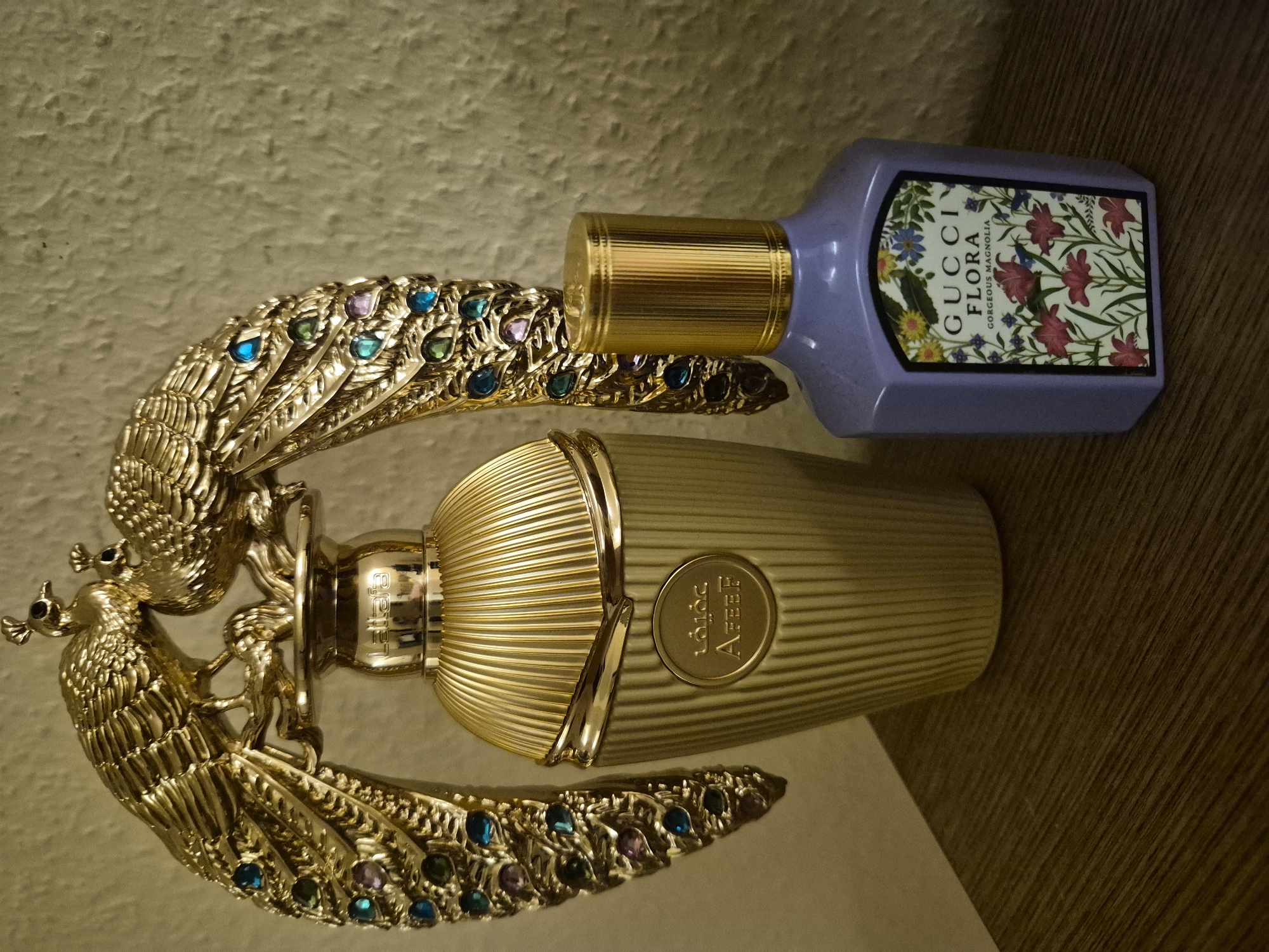 The image displays two perfume bottles on a textured surface, possibly a wall and a wooden table. One bottle is a light lavender color with a gold cap and features a label with botanical illustrations and the text "GUCCI FLORA GORGEOUS MAGNOLIA." This bottle is positioned horizontally. The other bottle is a striking gold with a ribbed design and a decorative cap. This bottle is partially obscured by a large, ornate gold peacock figurine with colorful gemstone accents in its feathers. The peacock figurine is positioned in front of the gold bottle, with its wings extending upwards and outwards, creating a dramatic visual. The overall presentation suggests a still life arrangement of luxury items. The lighting is warm, casting soft shadows, which could indicate indoor lighting. The scene appears static, with no activity taking place. The visible text on the lavender bottle clearly identifies the brand and fragrance. A circular emblem with Arabic-style script and the letters "AFEF" is visible on the gold bottle.
