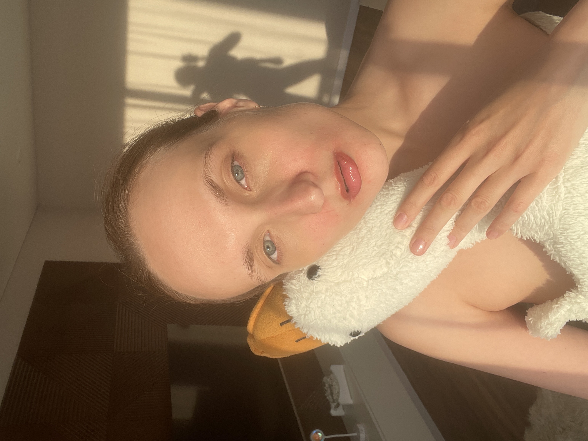 An angled, close-up shot captures a young person indoors, bathed in warm natural light. The subject, appearing to be female, has light skin, light blue-grey eyes, and light hair visible around her forehead. Her head is tilted, and she looks directly at the camera with slightly parted, glossy lips. She holds a white, fluffy plush toy, resembling a goose or duck with an orange beak, against her upper body.
Strong sunlight streams into the room, casting distinct shadows on a light-colored wall in the background, suggesting either morning or late afternoon. These shadows include patterns indicative of window blinds or a frame, and one prominent shadow resembles a human figure or an object. The setting is an indoor space, possibly a residence in Podshivalovo, Russia. A dark, textured surface, likely furniture, is visible in the lower left, and a reflective object rests on another dark surface in the bottom right. The image conveys a relaxed and casual mood, with no visible text.