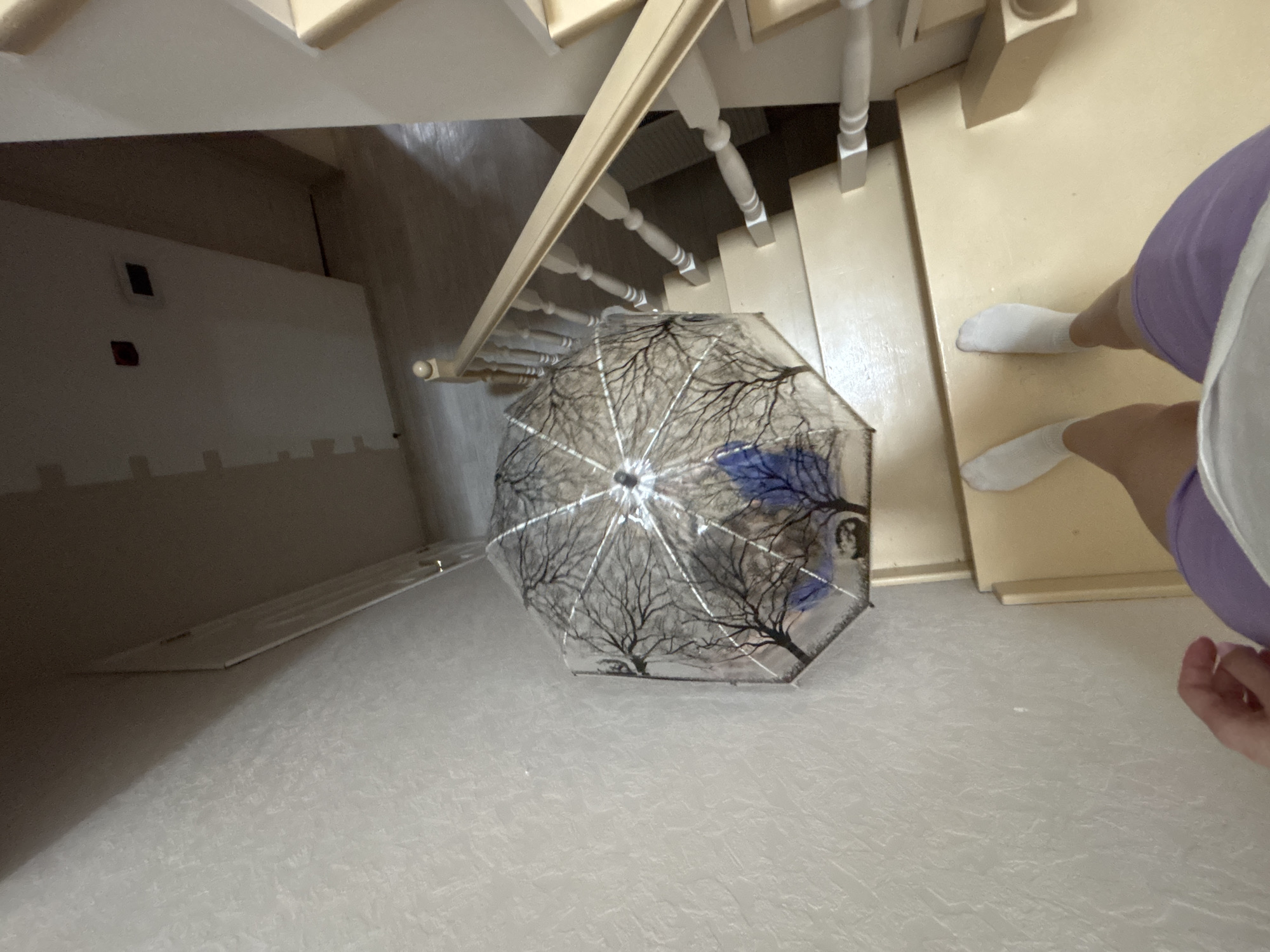 The image shows a view from above, likely from a staircase, in a location presumed to be Ферма, Russia. The floor is covered with a light, textured material. An umbrella with a design featuring tree branches is lying open on the floor. Part of a person's legs wearing white socks and purple clothing is visible on the right side of the image. The stairs are made of a light-colored material, potentially wood. The space under the stairs is visible, revealing a white wall with two small, dark squares on it. The overall lighting is bright.