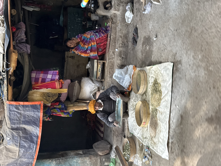An Indian storefront shows two individuals and goods for sale. One woman sits inside the doorway, with piles of textiles behind her. Outside, a man with a yellow turban is sorting seeds on a white sheet with three sieves. A variety of sorted seeds are visible. The area around the sheet is a weathered concrete surface. Overall, the scene conveys a small business or market operation.
