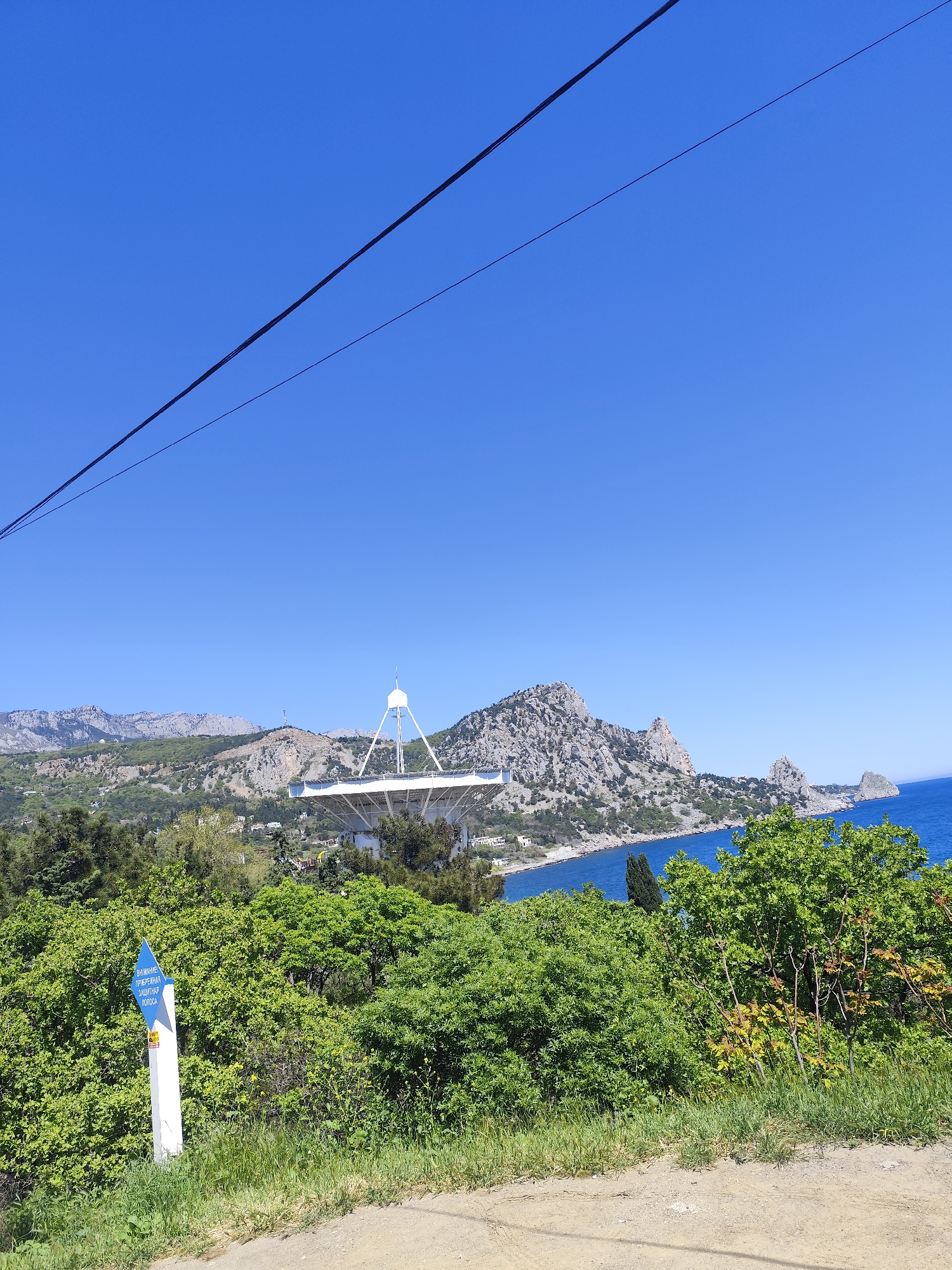 A landscape photograph shows a clear, blue sky over a coastline with a large, white radio telescope in the distance. The landscape features rolling hills covered in green vegetation, with a body of water visible on the right side of the frame. A blue sign stands near the roadside vegetation. The sky is a clear, uniform blue, with a black power line cutting across the upper left of the image.
