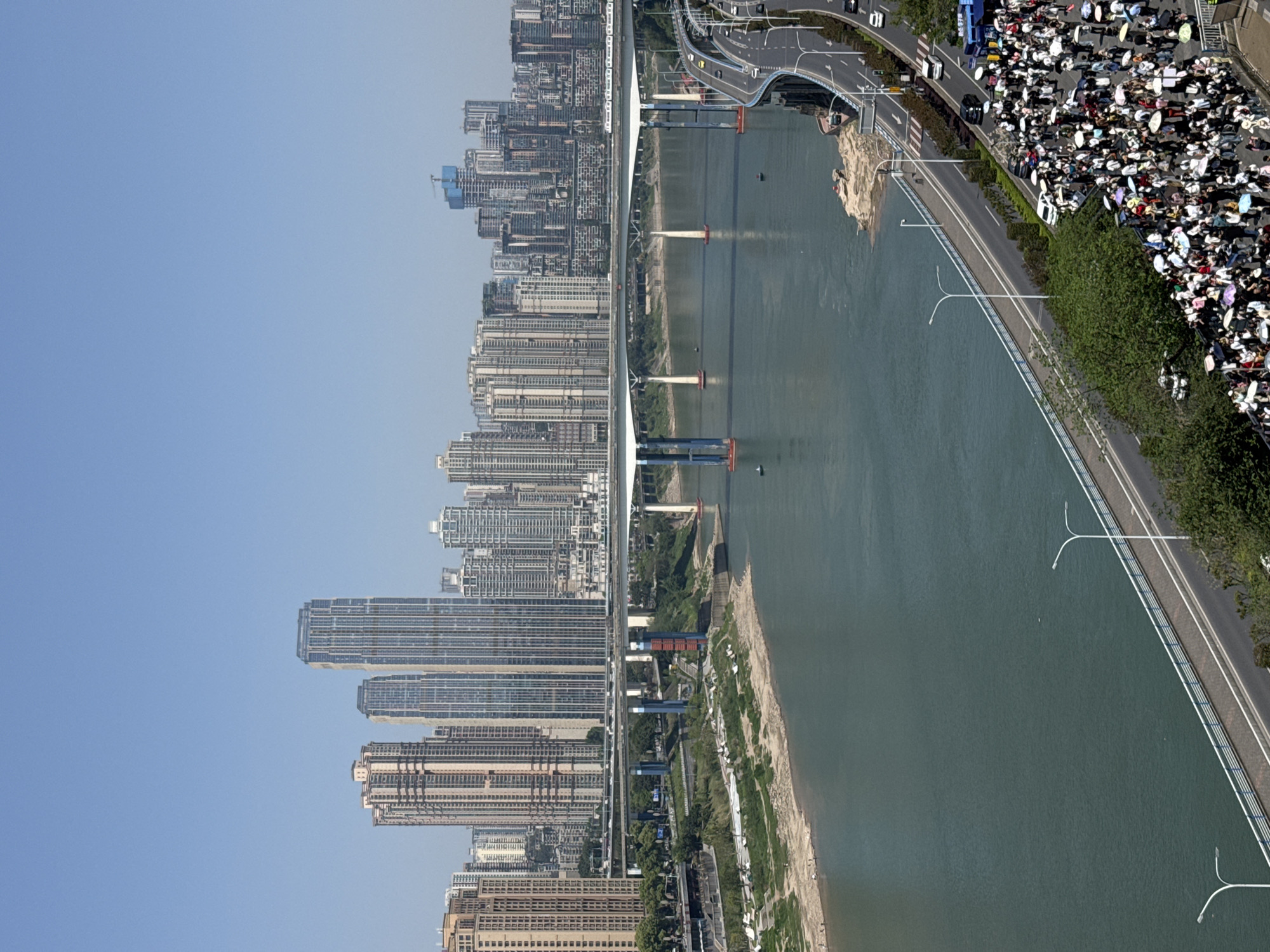 The image depicts an aerial view of the city of Yuzhong District, China, with a prominent body of water bisecting the scene. Parallel to the water, there is a densely populated area, suggesting a bustling urban environment. Buildings line the edge of the water, while a large crowd of people is gathered near the shore. Many individuals are holding umbrellas, indicating either sunny weather or protection from some falling debris. A road runs alongside the water, and cars can be seen driving on it. A bridge spans the waterway, connecting different parts of the city. The overall impression is a vibrant and active urban landscape.