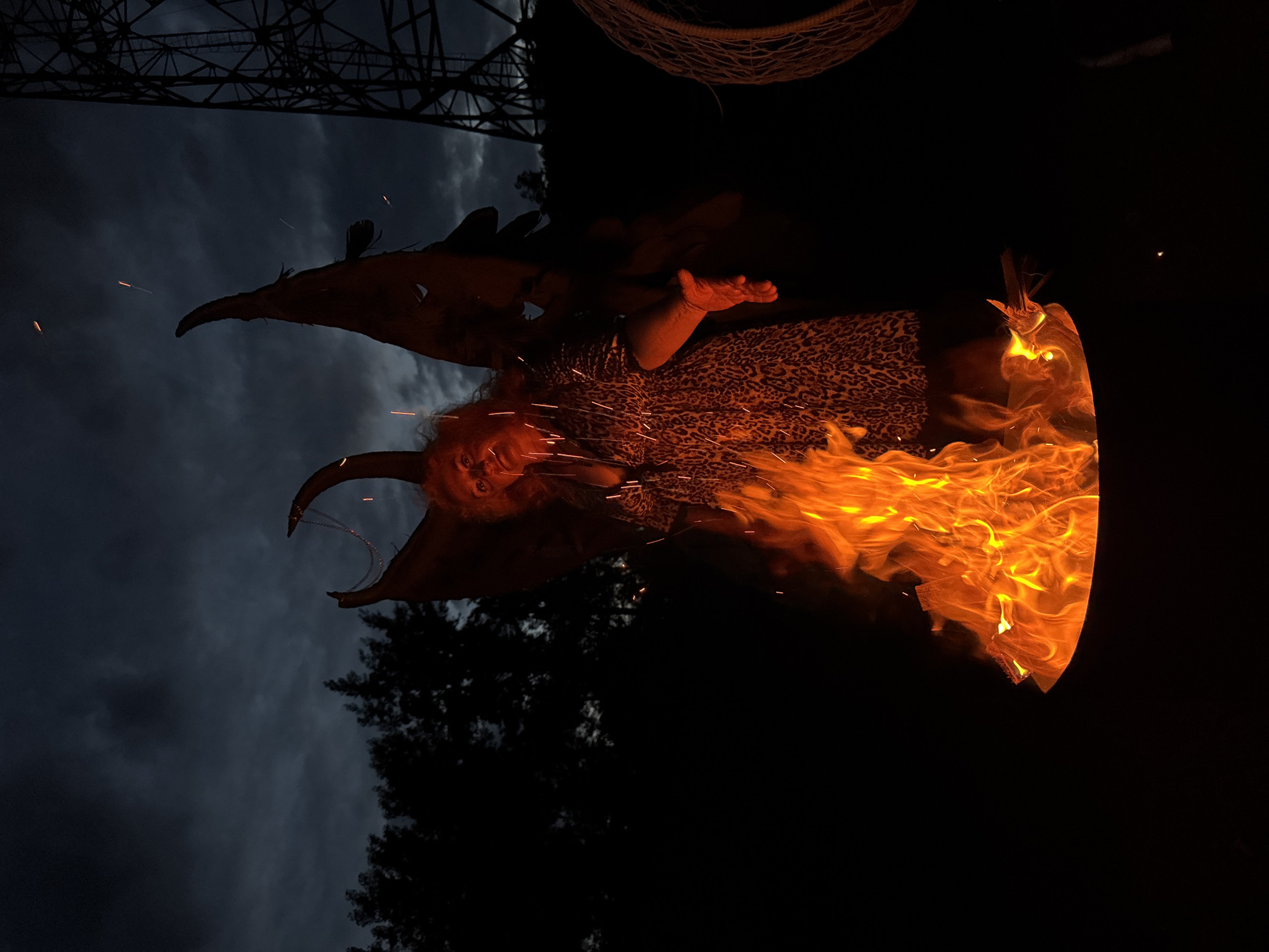 A person with dark, curly hair and fair skin, wearing a leopard print dress, stands behind a bonfire in a metal fire pit. They have large, dark, leathery wings attached to their back. Their face is illuminated by the fire, casting a red glow on their features. Their right hand is extended towards the viewer. Sparks are visible in the air. In the background, dark trees line a hill, and a tall metal tower is visible on the right. The sky is dark and cloudy, indicating it is evening or nighttime. The scene appears to be outdoors, possibly in a rural or park-like setting, given the presence of trees and the open sky. There is no discernible information within the image that could definitively place this scene in Chaikovsky, Russia.