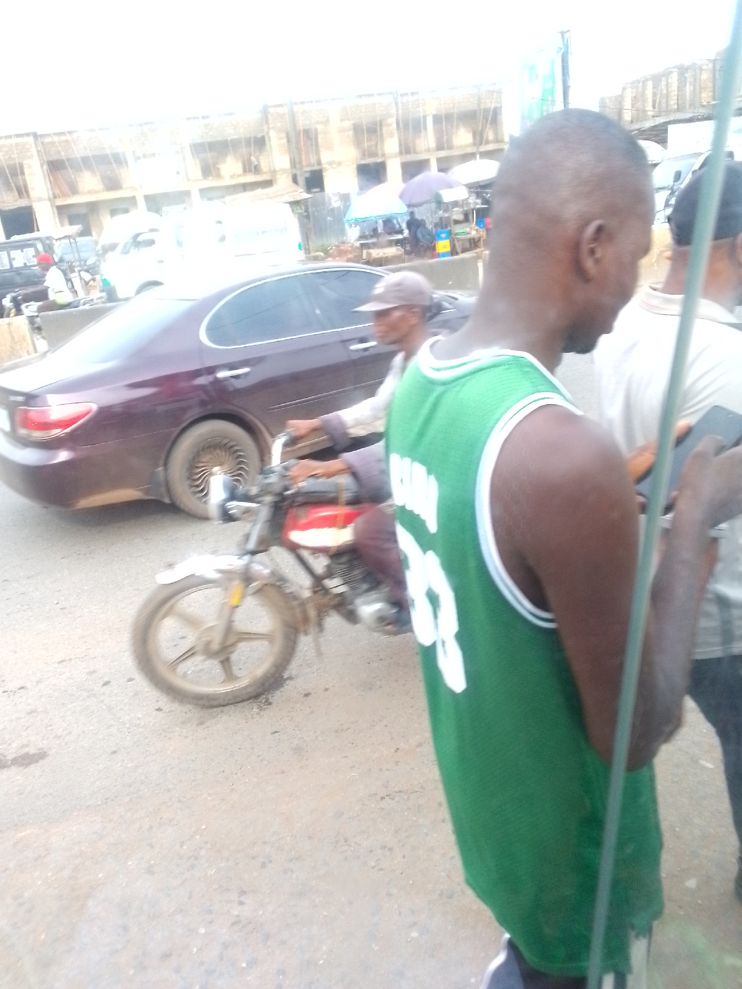 The image captures a street scene in Ihiala, Nigeria. A man in a green basketball jersey with white trim and the number 33 on the back stands in the foreground, partially obscuring the view. To the left, a motorcyclist with a baseball cap drives a red and silver motorcycle on the road. Behind the motorcycle, a maroon sedan is parked. In the background, there are other vehicles, stalls with umbrellas, and buildings. The road surface is asphalt and appears to be in a state of disrepair. Another man in a light-colored shirt stands to the right, looking at a phone.