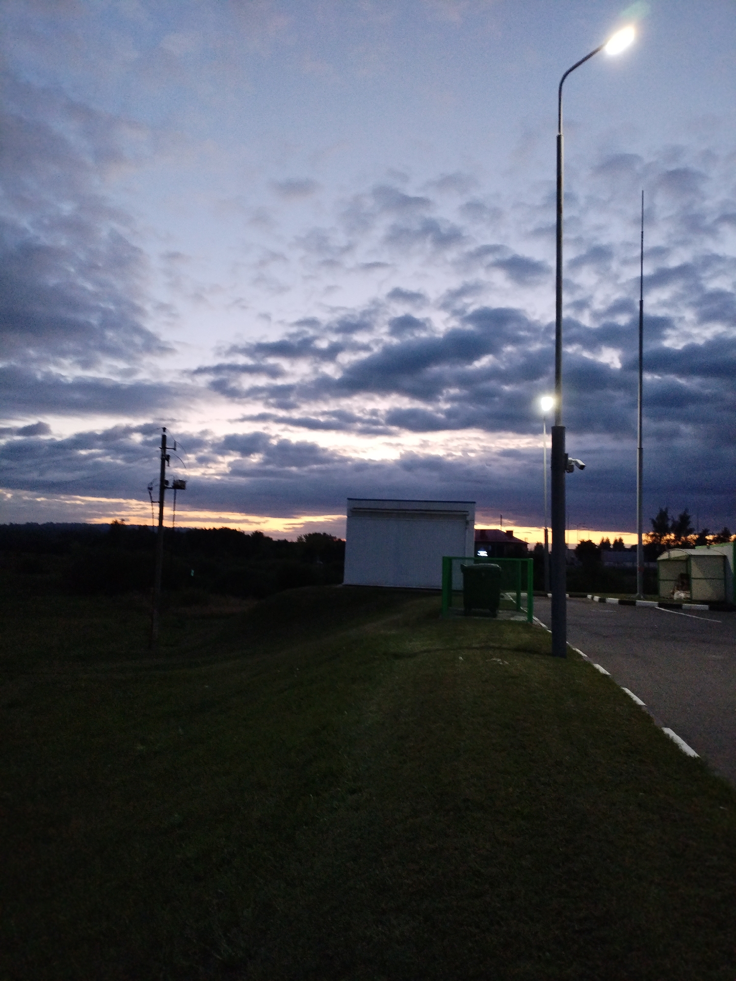 The image depicts a rural setting at dusk in Bialynichy, Belarus, characterized by a dramatic sky filled with dark, heavy clouds and streaks of orange and yellow light from the setting sun. In the foreground, a well-maintained grassy slope leads down to a dark expanse of land, likely fields or trees, extending to the horizon. A weathered utility pole with electrical equipment stands on the left side of the slope.
Further into the scene, a modern white rectangular building is situated on the right side of the slope, adjacent to a green metal fence enclosing a dark green trash receptacle. A paved road curves around the slope, bordered by white and black markings. A tall, slender streetlight with a bright illuminated head stands prominently on the right, casting a faint glow. A security camera is attached to a pole near the streetlight. Further down the road, another taller pole and a small, open-sided structure with a light-colored roof are visible. The overall atmosphere is tranquil, suggesting the end of a day in a less populated area.