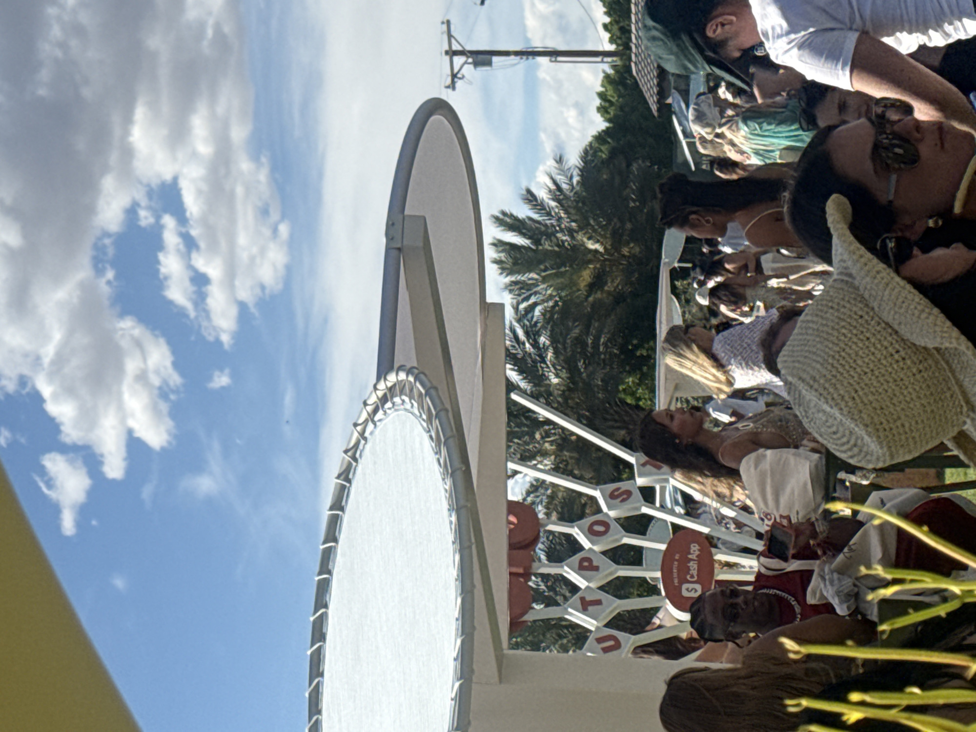 A medium shot shows a gathering of people under a structure in the city of Indio, California, United States. The structure includes a large, circular white surface and is labeled with the word "SPOT." The people are seated at tables and appear to be socializing and relaxing. One of the people is using a cell phone that is being used for Cash App payments. The environment includes palm trees, a blue sky, and clouds, suggesting an outdoor setting. A utility pole is also visible in the background.
