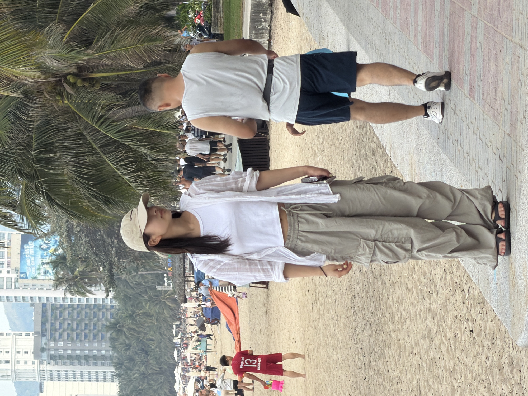 The image captures a bustling beach scene in Nha Trang, Vietnam, likely near Tran Phu Beach. In the foreground, a young woman wearing a beige baseball cap, a white crop top, a loose white sheer shirt, and baggy tan cargo pants stands facing left. She is wearing dark sandals. To her right, further down the paved walkway, a man with short hair and a white sleeveless shirt, dark shorts, and white athletic shoes is walking away from the camera. The background reveals a crowded beach with numerous people, beach umbrellas, palm trees, and tall buildings that are characteristic of Nha Trang's cityscape. The sand is light brown, and the paved walkway has a patterned surface. A child in a red shirt with the number 13 on it is visible on the sand to the left, holding a pink bucket and shovel. The overall atmosphere suggests a warm, sunny day at a popular tourist destination.