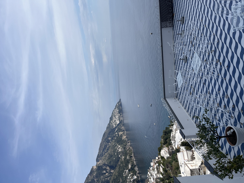 This high-angle, panoramic photograph captures a serene coastal scene in Positano, Italy, from an elevated outdoor terrace.
The upper portion of the image displays a light blue sky with scattered, wispy white clouds. Below the sky, the deep blue waters of the Mediterranean Sea extend to the horizon, appearing calm with minimal surface disturbance. Several small boats, white or light-colored, are visible on the water; one is actively creating a wake.
To the left, a steep, verdant mountainside descends towards the sea, densely populated with numerous white and light-colored buildings characteristic of Positano's terraced architecture. The coastline curves gently to the right.
The foreground is dominated by a large, unoccupied outdoor terrace. The terrace floor is tiled with a distinctive wavy pattern of alternating dark blue and white stripes. Numerous sets of white, wrought-iron furniture, consisting of square glass-topped tables and matching chairs, are arranged geometrically across the patterned tiles. A white concrete parapet with decorative white metal railings, featuring some black wrought-iron detailing on the right, borders the terrace. In the lower left corner, a potted plant with green leaves, possibly a small citrus tree, sits on the terrace floor. No individuals are present in the scene. The overall lighting suggests daytime conditions.