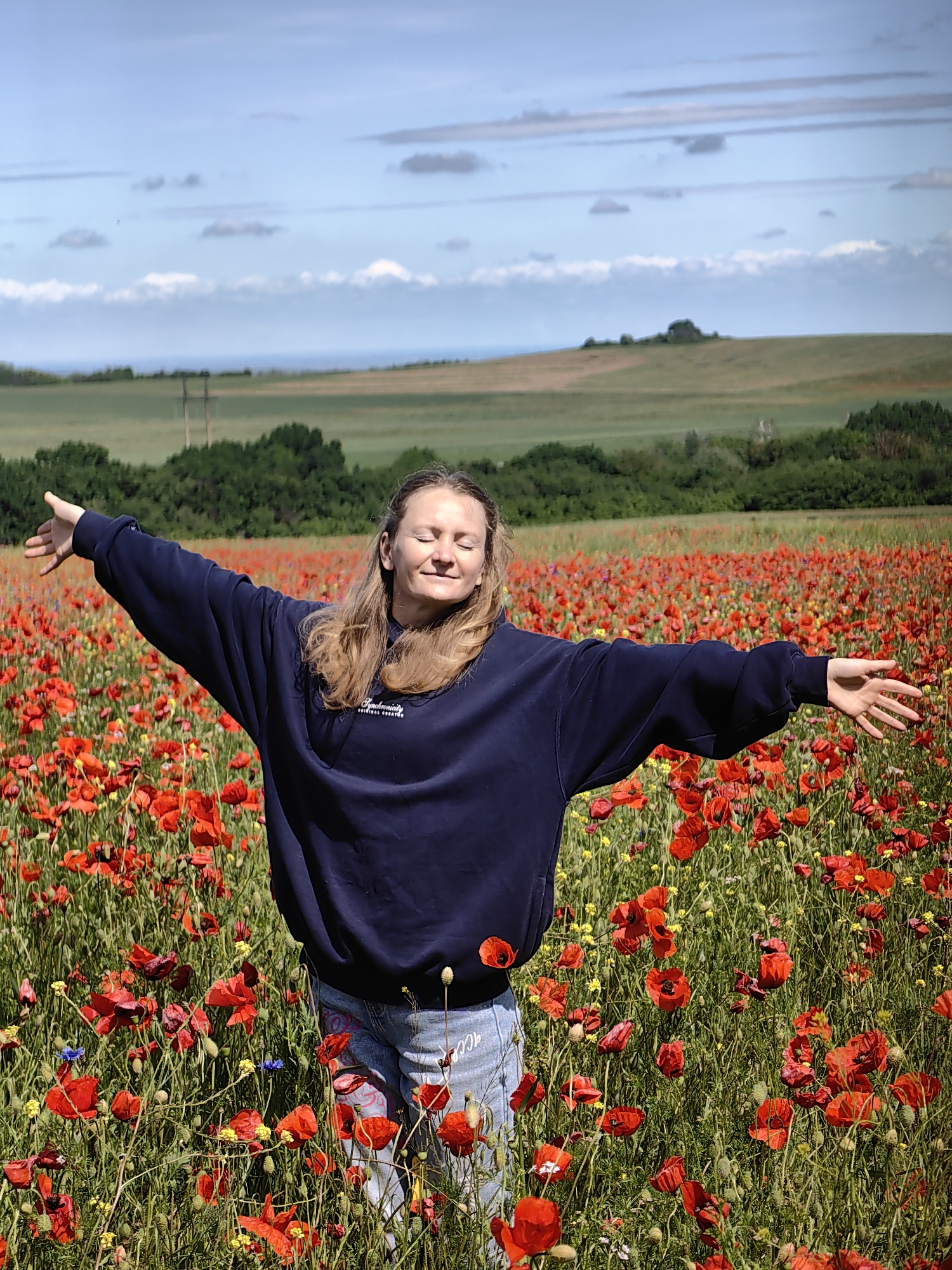 A woman stands in a field of red poppies under a partly cloudy sky. She is wearing a dark blue sweatshirt with the words "Synchronicity ORIGINAL CREATIVE" on the chest, and jeans with drawings on them. Her arms are outstretched. In the background are green fields and trees. The location is consistent with Ароматновское сельское поселение, Ukraine, given the poppy fields and landscape.