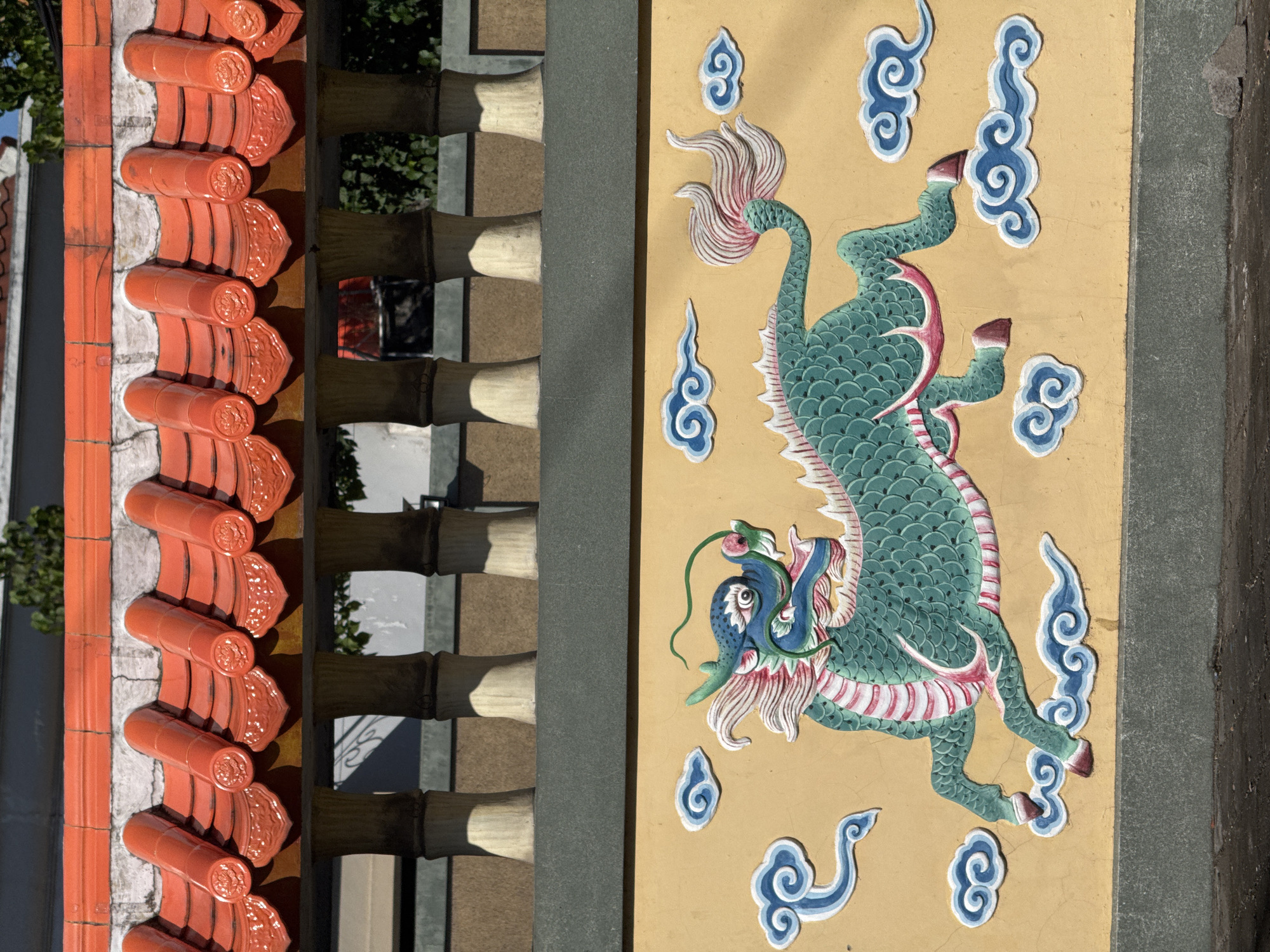 This image depicts a section of a building adorned with traditional Balinese architectural elements. On the left, there's a series of orange, barrel-shaped roof tiles arranged in a row. Adjacent to the tiles is a structure composed of stacked, horizontally aligned bamboo-like elements. A grey, flat wall separates these elements from a yellow wall featuring a prominent dragon-like creature in relief. The creature is teal with pink and white accents and is surrounded by stylized blue clouds. The building is located in Denpasar, Indonesia.