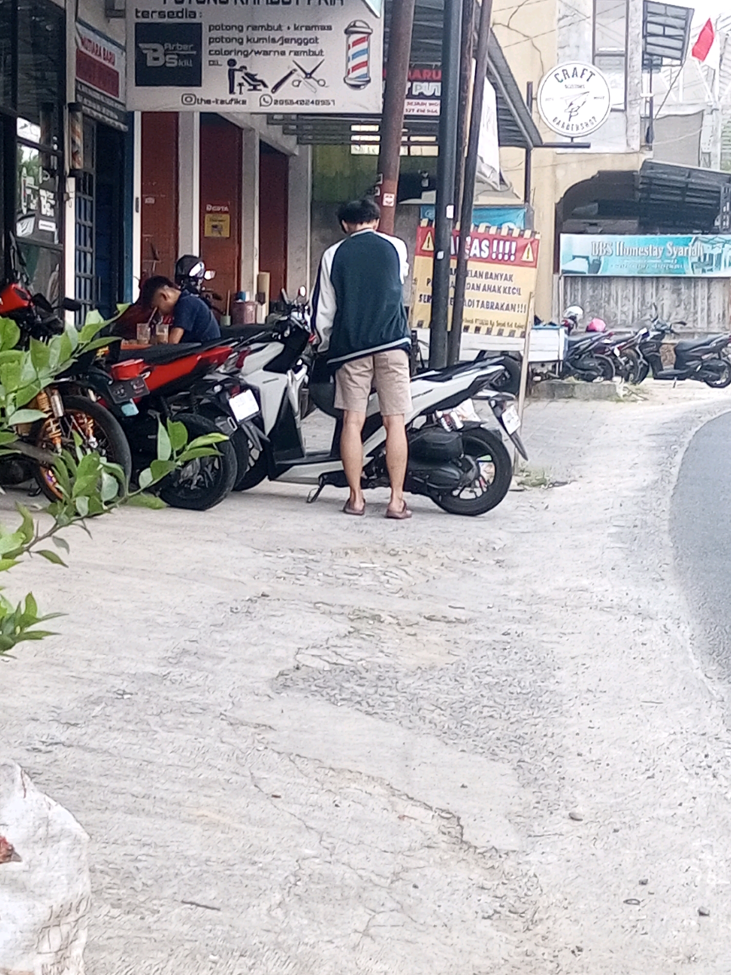 The image depicts a street scene in Mojotengah, Indonesia, likely near a barber shop and an accommodation service. In the foreground, several motorcycles are parked on a concrete sidewalk. To the left, a person is seated at a table with drinks, appearing to be socializing. A man is standing on the sidewalk, wearing shorts and a jacket, facing away from the camera. Behind him, the facade of a building with signs for a barber shop and a homestay is visible. The barber shop sign features a barber pole graphic and text in Indonesian, indicating services like haircuts, coloring, and hair treatment. The homestay sign, in blue and white, reads "DBS Homestay Syariah." The overall environment suggests a commercial area with a mix of services.