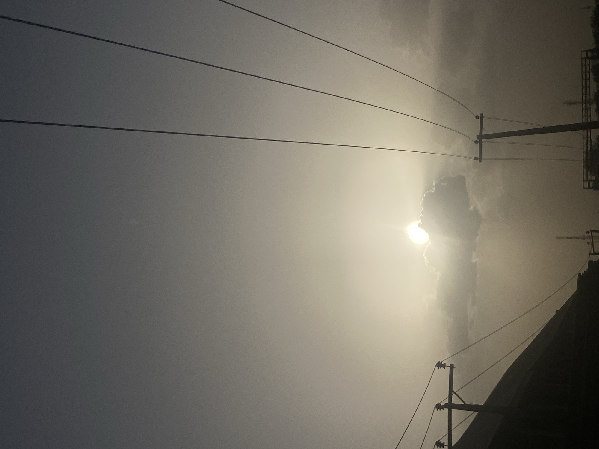 The image shows a sky view in Tudun Wada, Nigeria. Power lines are visible, crisscrossing the sky. The sun is visible through a thin layer of clouds. Power poles and a building are visible at the bottom right of the frame.