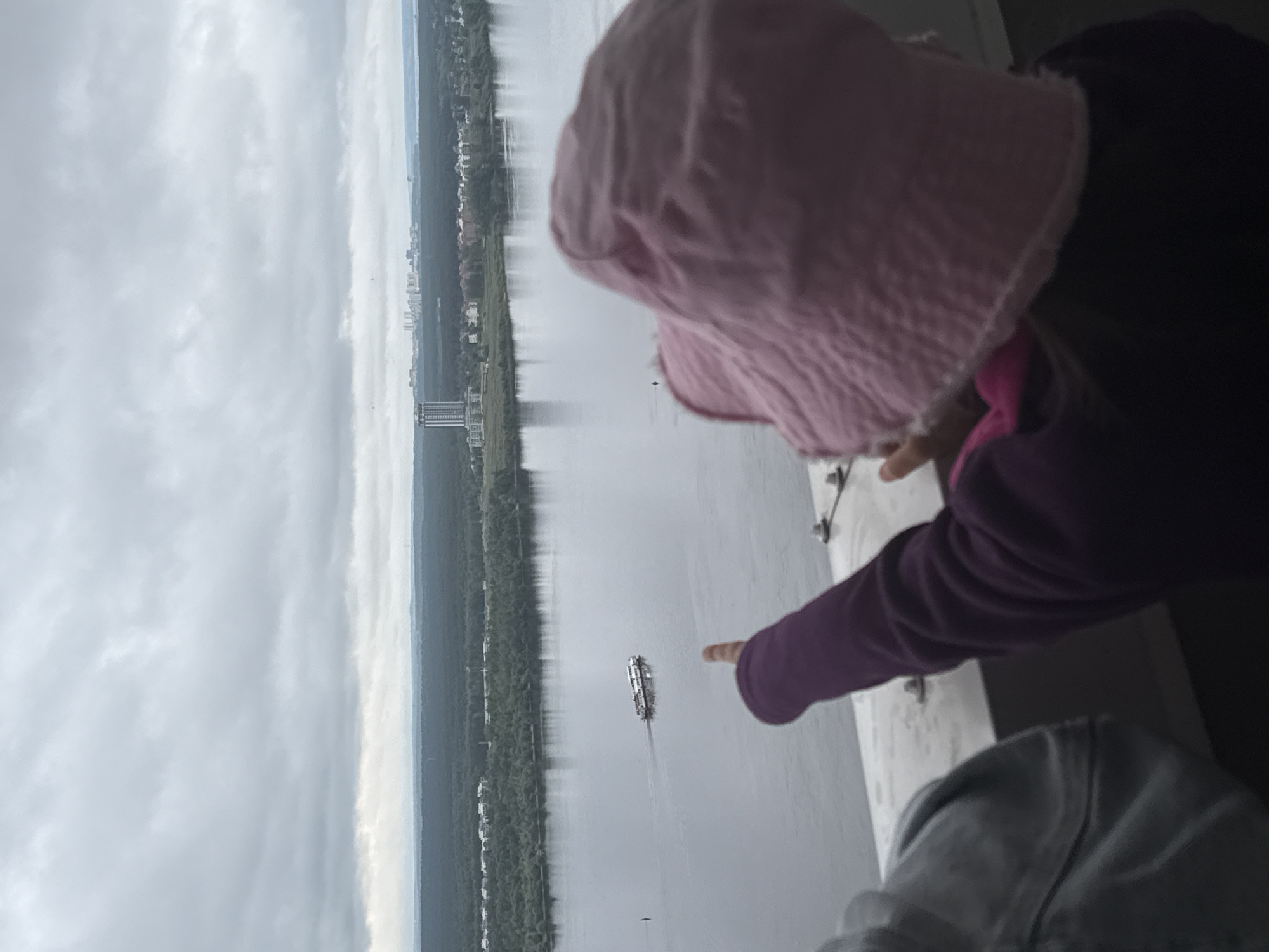 From an elevated perspective in Perm, Russia, the image captures a wide river under a heavily overcast sky. In the foreground, the back of a person wearing a pink bucket hat and a dark purple long-sleeved top is visible, with their right arm extended and index finger pointing towards the river. A grey garment is partially visible on the far left. The person is situated behind a light-colored metal railing or ledge secured by bolts.
The midground features a broad river, appearing calm and reflecting the grey sky. A white and grey passenger boat with multiple decks is observed moving from left to right, leaving a distinct wake. A dark, slender marker or buoy stands vertically in the water to the left of the boat. Both banks of the river are densely forested with dark green trees, interspersed with scattered residential or commercial structures.
In the background, a prominent tall, multi-story building with a distinctive top stands on the far bank, casting a reflection in the river. Further in the distance, a cluster of high-rise buildings forms a cityscape silhouette against the horizon. The entire sky is uniformly covered with thick, grey clouds, indicating an overcast day.