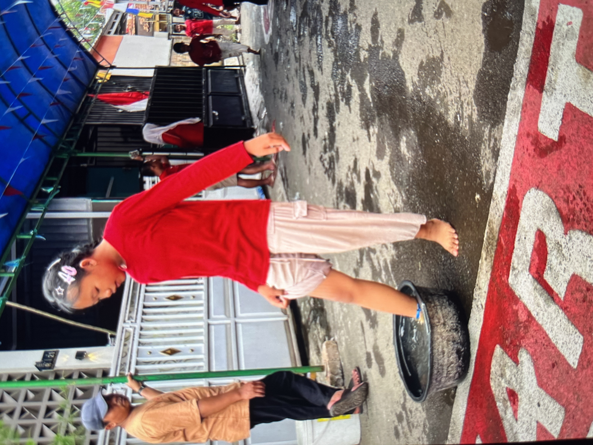 In the city of Cirahayu, Indonesia, a young girl with her hair in a braid, adorned with star-shaped and "40" hair clips, stands barefoot on a wet, paved surface. She is wearing a red long-sleeved shirt and light-colored, loose-fitting pants, with one leg submerged in a black plastic basin filled with water. The girl is in the process of washing her foot. Beside her, to the left, a man wearing a blue baseball cap and an orange t-shirt stands with his right hand on a green pole. He is looking towards the girl. Behind them, a white fence with diamond-shaped details separates the paved area from what appears to be a building. The background shows more buildings and people, with Indonesian flags visible, suggesting a celebratory or community event. The ground is wet and reflects the surrounding environment, indicating recent rain or spilled water. The scene is outdoors, under a blue awning decorated with small triangular flags. Red and white painted markings are visible on the ground in the foreground.