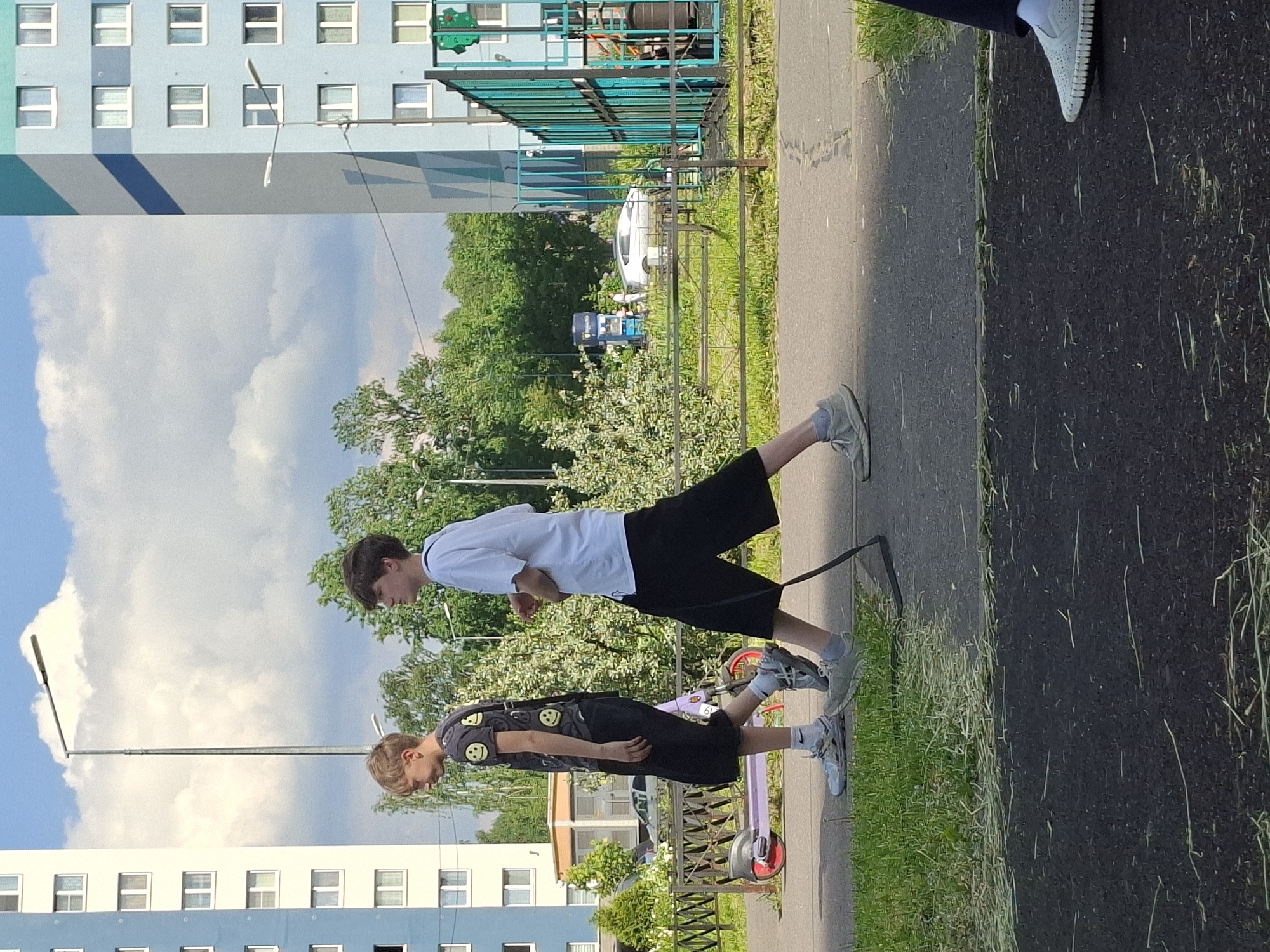 The image captures two males, an older youth and a younger boy, standing on a paved pathway next to a grassy area under bright daylight. The sky is partly cloudy with large white clouds against a blue background. The scene is likely an urban residential area or a park in Saint Petersburg, Russia.
The older male, positioned on the left, wears a white short-sleeved t-shirt, dark shorts, and light-colored sneakers with white socks. He has dark hair and is looking downwards. He appears to be holding a thin, dark leash or rope that extends towards the lower center of the frame.
To his right stands a younger boy, who is next to a purple scooter with red wheels. The boy wears a dark t-shirt decorated with multiple yellow smiley face emojis, dark shorts, and light-colored sneakers with white socks. He has lighter brown hair and is also looking down. The scooter is upright on the path beside him.
In the background, light blue multi-story residential buildings with white window frames are prominent on the left. Green trees and dense foliage line the right side of the path, separated by a green metal fence. Beyond the fence and trees, vehicles, including a white van, are faintly visible. A street lamp extends into the sky from the left side of the frame.
The individuals appear to be paused during an outdoor activity, possibly a walk or a break from scootering. The weather is sunny, indicating a fair-weather day. No specific emotions are clearly discernible from their expressions. The only visible text is the pattern of yellow smiley faces on the younger boy's t-shirt.