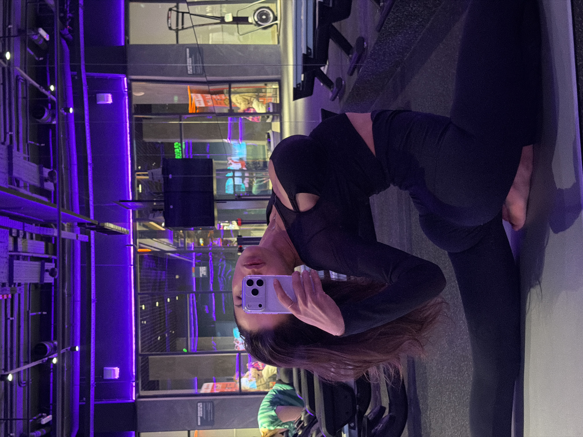 A woman in a black athletic outfit is taking a selfie with a lavender phone in a fitness center. She is standing on a gray yoga mat near a wall with floor-to-ceiling windows that reflect an urban environment. The reflections suggest an area with bright neon lights, possibly a commercial district, with purple and other colored lights. There are hints of machinery and architectural details in the reflections. Other people can be seen in the background. While the location in Krasnoyarsk, Russia, is stated in the prompt, there are no distinguishing features in the image itself to confirm this claim.