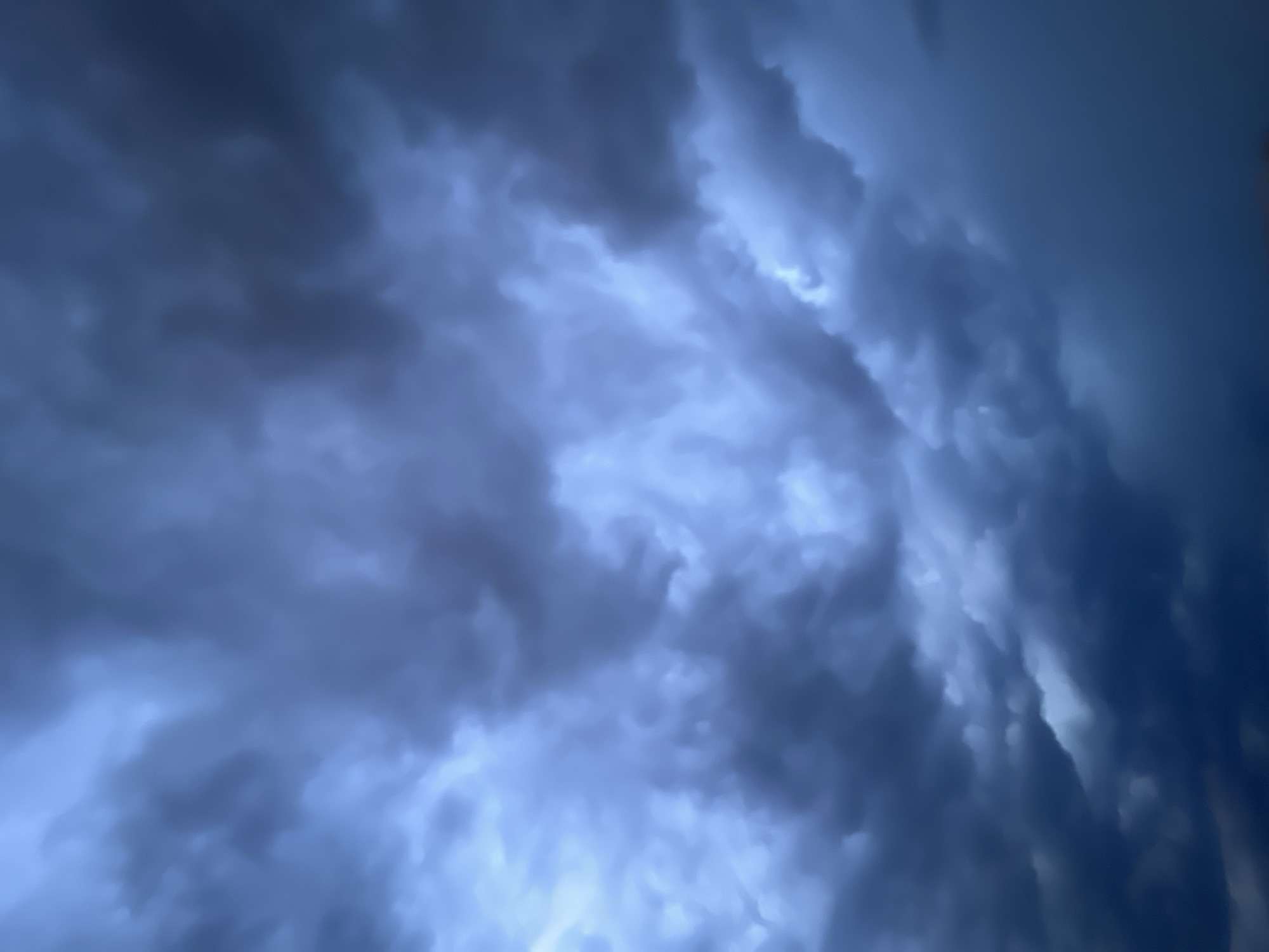This is a vertically oriented, full-frame, close-up photograph of a turbulent sky filled with dark, heavy storm clouds. The clouds exhibit a deep blue-grey hue with variations in tone, suggesting depth and density. Light appears to be breaking through in certain areas, creating brighter, almost ethereal patches that contrast with the surrounding darkness. The overall impression is one of an impending storm, with dramatic and imposing cloud formations dominating the entire frame. There is no evidence of the city of Đà Nẵng, Vietnam, or any other environmental or human elements in the image.