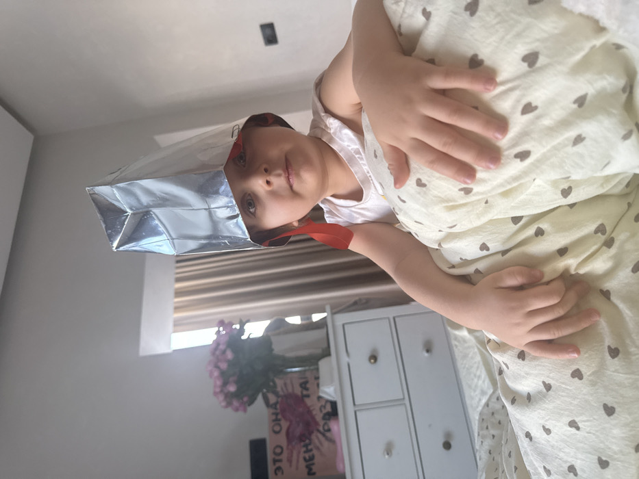 An indoor residential scene in Khimki, Russia, features a young child, approximately 2-5 years old, seated upright on a bed. The child, with fair skin and dark eyes, wears a light-colored sleeveless top and a makeshift helmet constructed from reflective, silvery material, secured with red fabric strips and paper clips. The child maintains a neutral expression, looking directly forward, with hands resting on a light-colored duvet patterned with small brown hearts. To the left, a white three-drawer dresser with light-colored knobs holds a clear vase with pink roses, a white rectangular object, and a sign with a red heart and Russian text, partially visible as "ЭТО ОНА МЕНЯ ТАК РАЗУ...". A window with light-colored curtains is visible behind the dresser. The walls are a neutral light color, and a dark electrical outlet or switch is present on the wall to the right. No specific identifying details within Khimki, Russia, are discernible.
