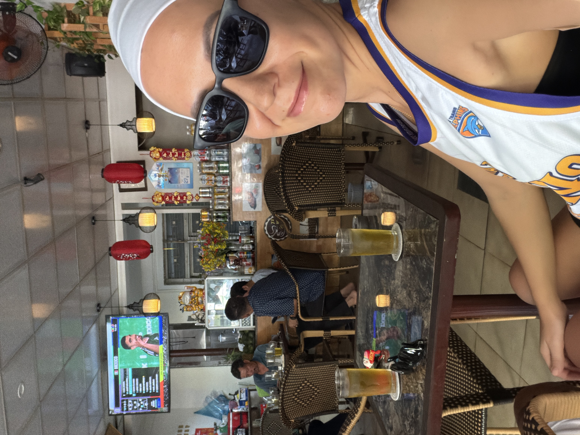 A woman, wearing a white headscarf and sunglasses, takes a selfie in a brightly lit indoor setting, possibly a cafe or restaurant, with a street scene visible in the background. She is wearing a white basketball jersey with purple and orange trim and a dolphin logo. The table in front of her has two glasses of iced beverages, small candles, and a black figurine. Behind her, other patrons are seated at tables, and a television displays a soccer match. Red lanterns hang from the ceiling, and shelves are stocked with drinks and decorative items, suggesting a location in Tay Nha Trang Ward, Vietnam.