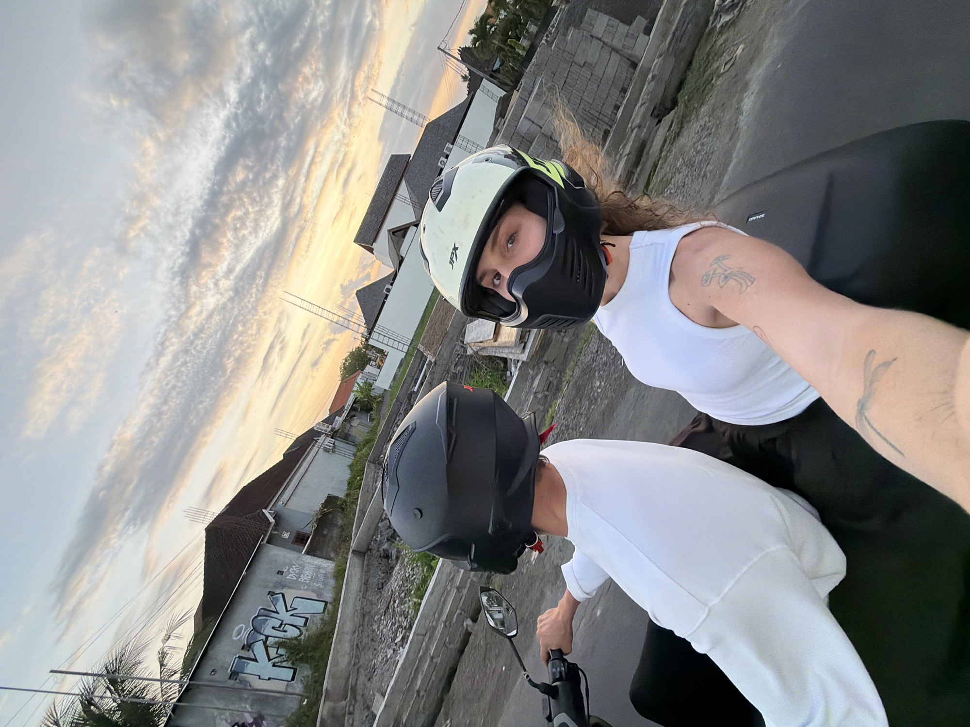 Two people, one in the foreground and one in the background, are riding a motorcycle on a road in North Kuta, Indonesia. The person in the foreground is wearing a black helmet and a white long-sleeved shirt. They are holding onto the handlebars of the motorcycle and looking at a mirror. The person in the background is wearing a white and black helmet and a white tank top. They have long, curly brown hair and a tattoo on their right arm.
The scene appears to be set at sunset, with a cloudy sky painted in hues of orange, pink, and gray. The background shows a street with buildings, some of which have graffiti on them. The overall atmosphere is casual and adventurous. The time of day is likely late afternoon or early evening, indicated by the sunset sky. The weather seems pleasant and conducive to riding a motorcycle.