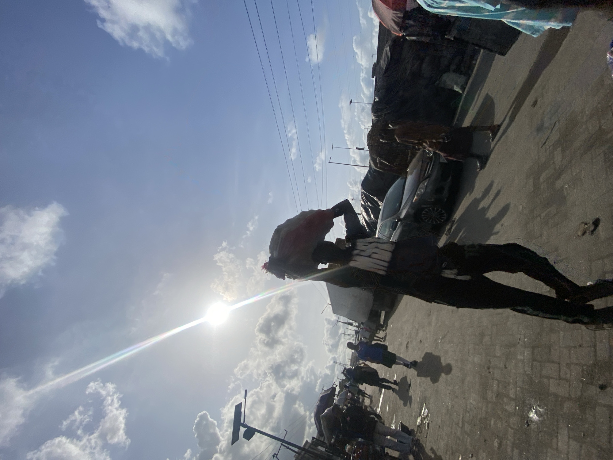 The image shows an outdoor scene in what appears to be a bustling urban environment. The sky is partly cloudy with the sun shining brightly. Several individuals are walking and standing around, suggesting a public space like a market or a busy street. There are vehicles parked along the side, including what appears to be a dark-colored car. Power lines stretch across the sky. Based on the mention of Lekki, Nigeria, it is plausible that the image captures a scene in a market or commercial area within the city, characterized by vendors, shoppers, and general activity. The perspective is slightly tilted, adding to the dynamic feel of the image.