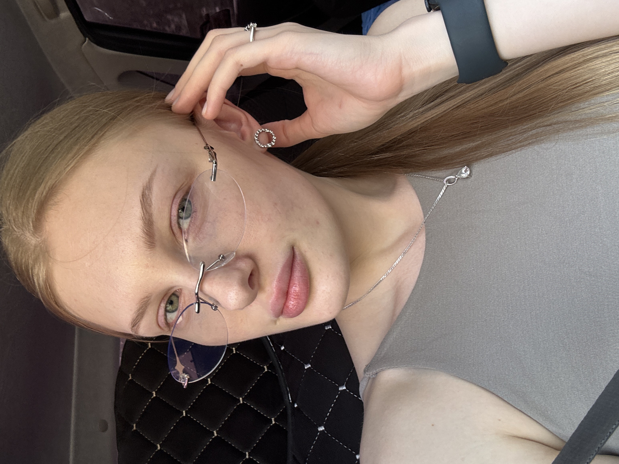 A woman is indoors, possibly in a car given the quilted pattern of the seat and partial view of a car window. She has blonde hair and is wearing rimless glasses, a silver necklace, a ring, a watch, and circular earrings. Her hand is near her ear, possibly adjusting the earring or hair. The image is a close-up, focused on the woman's face and upper body. There is no clear information to establish that this picture was taken in Perm, Russia.