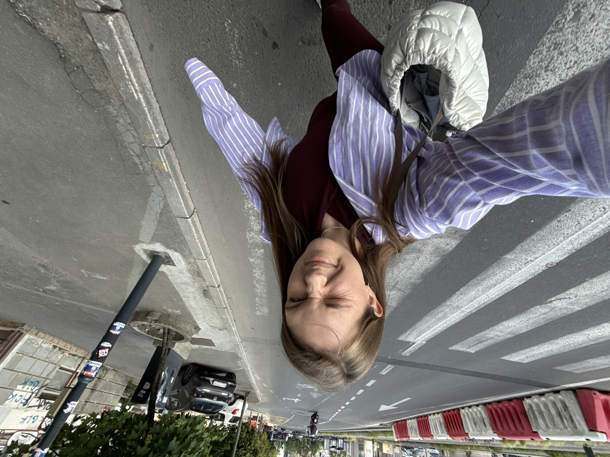 A low-angle, eye-level image of a street scene. A young woman is positioned prominently in the foreground, her face close to the camera and squinting. She wears a striped purple and white button-down shirt over a maroon top and is carrying a puffy white bag. The background shows a city street with a crosswalk, a street with a visible arrow lane marking, a parked black car, road dividers, a lamppost with stickers, and some greenery. Distant buildings are visible in the background. Based on available evidence, the location is Belgrade, Serbia.