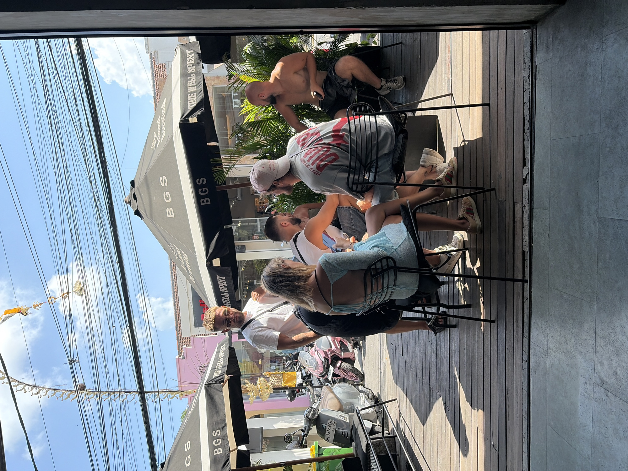 The image depicts an outdoor seating area on a wooden deck, likely at a cafe or restaurant in North Kuta, Indonesia. Several people are seated at tables beneath large black umbrellas that bear the branding "BGS" and "WELL SPENT". In the foreground, a woman wearing a light blue dress is seated, facing away from the camera. To her left, a man in a white t-shirt and black shorts stands, looking towards the seated group. Further back, other individuals are visible, some seated, some standing. In the background, a street scene is apparent with parked scooters, including a prominent pink scooter. Overhead, a complex network of electrical wires is strung across a bright blue sky with scattered clouds. The overall atmosphere appears casual and relaxed, consistent with a warm, sunny day.