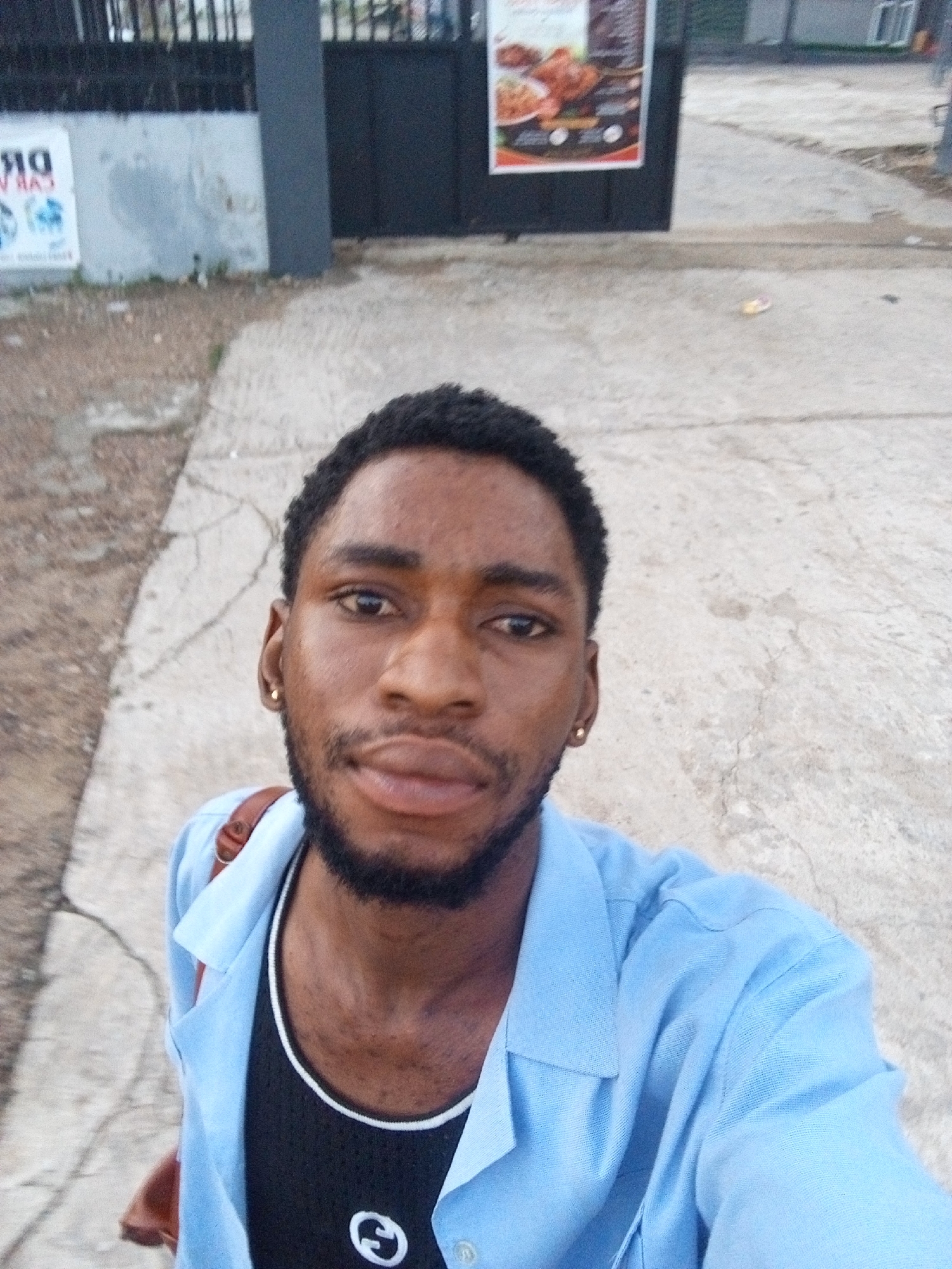 A young Black man with short black hair, a beard, and a gold earring in his left ear takes a selfie outdoors. He is wearing a light blue collared shirt over a black tank top with a white circular logo. A brown strap from a bag is visible over his right shoulder. He is standing on a paved surface, possibly a sidewalk or pathway, with cracks in the concrete. In the background, a black metal gate with vertical bars is visible, as well as a white wall with some graffiti or posters. To the left of the gate, there's a poster with food images. The environment appears to be in an urban setting in Lokoja, Nigeria, with elements suggesting a commercial or public area.
