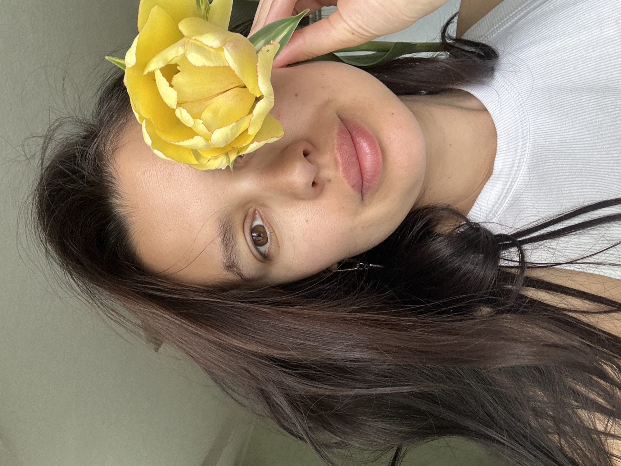 A woman is seen close-up, with a yellow flower held near her face, obscuring her left eye. She has brown hair, light skin, and brown eyes. She's wearing a white t-shirt. The background is a plain, light color. The location is not discernible from the image.