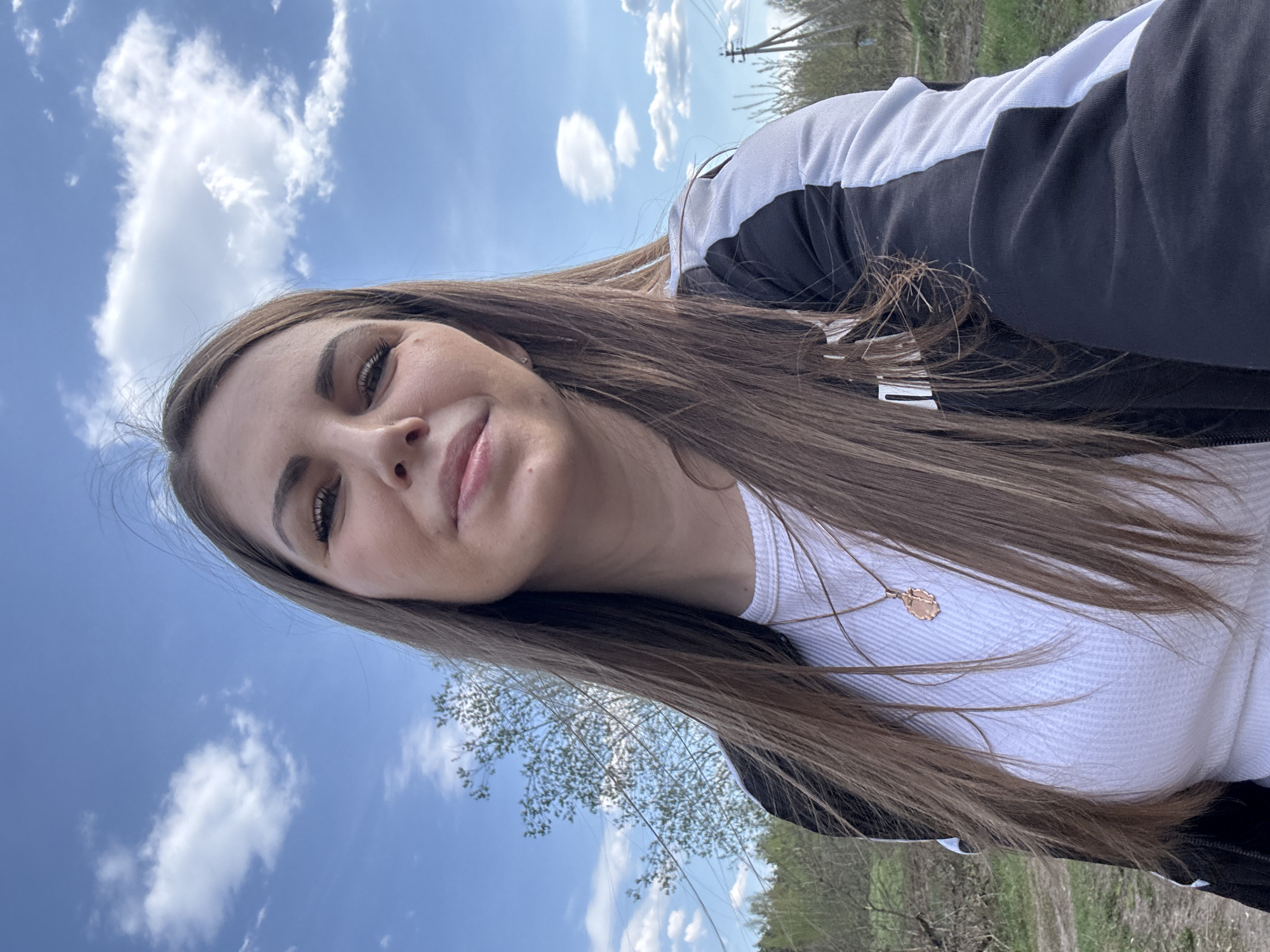 A young woman with long brown hair poses in a selfie. She wears a white top and a black and white jacket. A gold necklace with a pendant is visible. The background shows a blue sky with white clouds. The landscape includes trees and a dirt path, suggesting a rural or semi-rural environment. The location is likely Раздолье, Russia, based on the query.