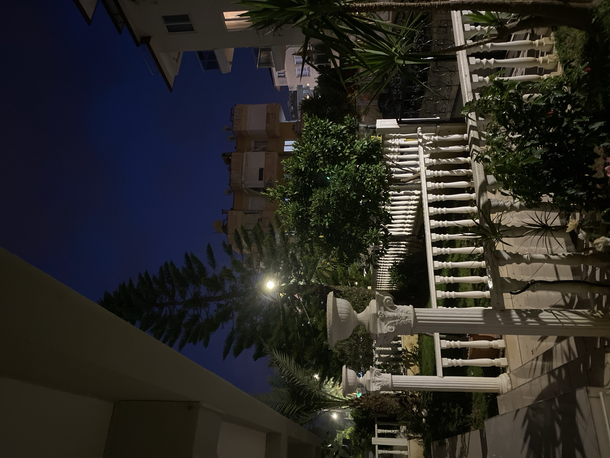 The image depicts an outdoor scene at night in Alanya, Turkey. Several multi-story buildings are visible in the background, with lighted windows suggesting residential or commercial occupancy. A dark blue sky is present overhead.
A white ornate balustrade dominates the foreground, framing a set of steps. Decorative white columns with elaborate designs stand at intervals along the balustrade. Green foliage and trees are interspersed throughout the scene, obscuring some of the architecture and providing a natural element. The lighting is dim, with a light source illuminating the trees and casting shadows on the balustrade and ground. The scene appears to be in a garden or courtyard setting.