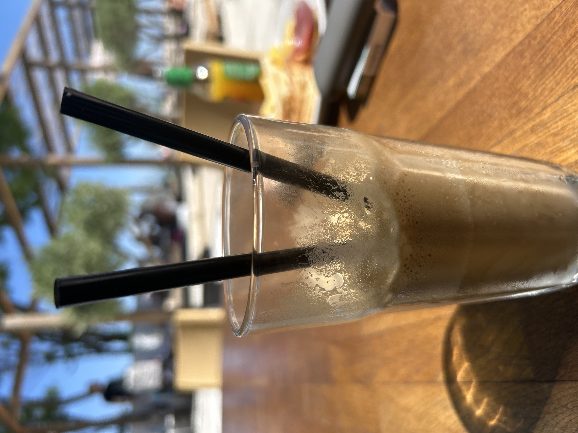 The image shows a close-up of a tall glass filled with a frothy, brown beverage, likely an iced coffee or a milkshake. Two black straws are inserted into the drink. The glass is sweating, indicating it's cold. The drink is placed on a wooden table. In the blurred background, there's a hint of an outdoor setting, possibly a cafe or restaurant with a wooden pergola structure overhead, green trees, and a blue sky. There are some out-of-focus figures and objects in the distance, including what appears to be a bottle and a plate of food. This scene is consistent with a casual outdoor dining experience at a cafe located in Burgas, Bulgaria, a city known for its coastal resorts and vibrant atmosphere, especially during warmer months.