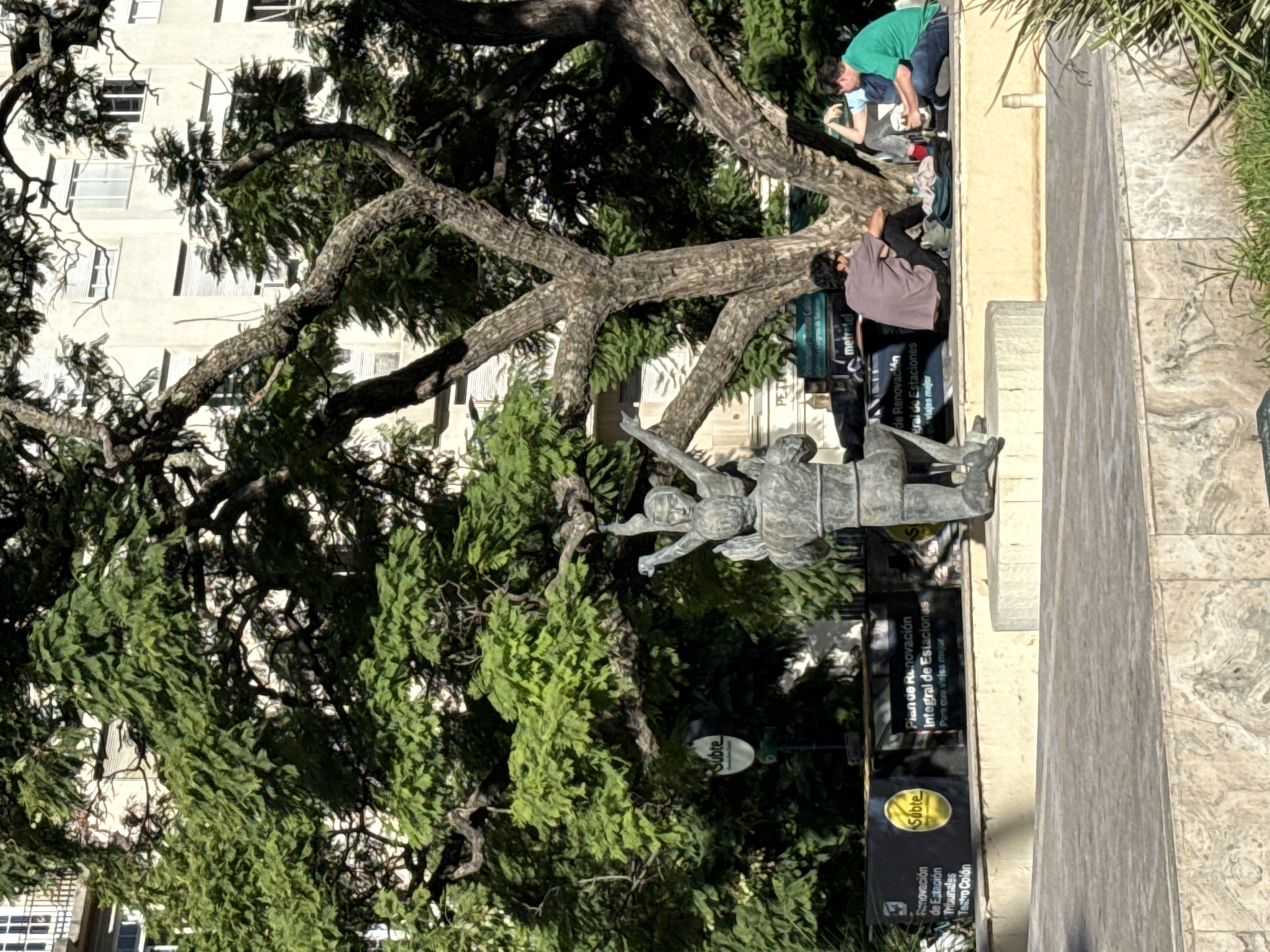This media file captures an outdoor urban scene in Buenos Aires, Argentina, featuring a large, gnarled tree, a metal statue, and several people under bright daylight.
A massive tree with thick, winding branches and dense green foliage occupies the left side of the frame, its rugged bark clearly visible. In the mid-ground, a dark metal statue depicts two figures in a dynamic pose, possibly embracing or reaching upwards, with their arms extended.
Several individuals are present: one person in a green shirt and blue pants is seen near the tree's base, seemingly engaged with something on the ground, while another in a light blue shirt is nearby. Further to the right, a person wearing a dark brown/purple shirt and dark pants is seated on a low, beige wall or bench. In the background, a light-colored building with multiple windows is visible through the tree's canopy. The ground is a mix of paved concrete and patterned tiles.
The scene is illuminated by strong sunlight, suggesting it's midday or afternoon. Visible text on signs includes: "Subte", "Renovación de Estación Tribunales Teatro Colón", "Plan de Renovación Integral de Estaciones Para que viajes mejor", and "metrotel", along with partial text like "de Renovación gral de Estaciones viajes mejor". These signs indicate the location is near a Buenos Aires metro (Subte) station undergoing renovation, specifically mentioning the Tribunales and Teatro Colón stations.