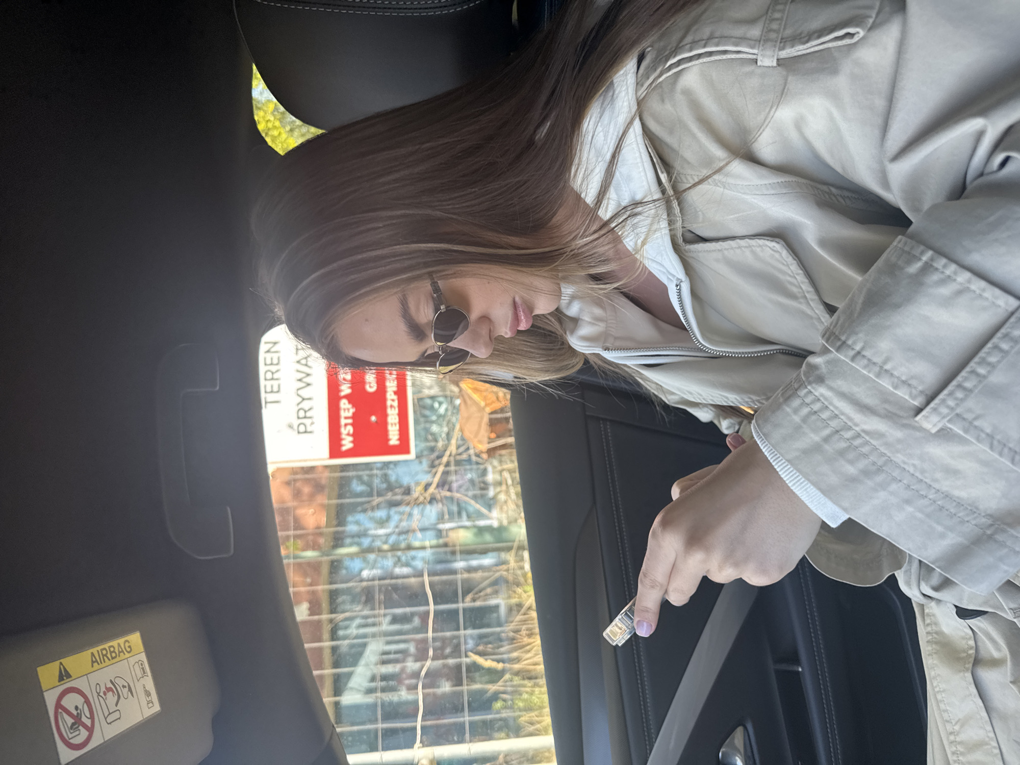 The image depicts a young woman seated in the passenger seat of a car, captured in a close-up, slightly angled view. She has long, light brown hair, is wearing sunglasses, and is dressed in a light-colored (possibly beige or cream) jacket or trench coat over a white top. Her head is tilted downwards, and her gaze is directed towards her right hand, which holds a small, transparent, possibly electronic device, near the car's seatbelt buckle. Her fingernails are painted a light lavender color.
The car's interior is visible, featuring dark upholstery on the seat and door panel, along with a seatbelt. A gray sun visor in the upper left corner displays a yellow "AIRBAG" warning label with pictograms, indicating safety information for the passenger.
Through the car window, an outdoor scene is visible, illuminated by bright daylight. There are hints of trees or foliage and a grid-like structure, possibly a fence or part of a building. A prominent red sign with white Polish text is clearly visible outside. The text reads: "TEREN PRYWATNY" (Private Property), "WSTĘP WZBRONIONY" (Entry Forbidden), and "GROZI NIEBEZPIECZEŃSTWEM" (Danger Threatens).
The overall impression is a casual, everyday moment of a woman inside a car, with the visible sign suggesting a location with restricted access, possibly in Warsaw, Poland, as per the context.