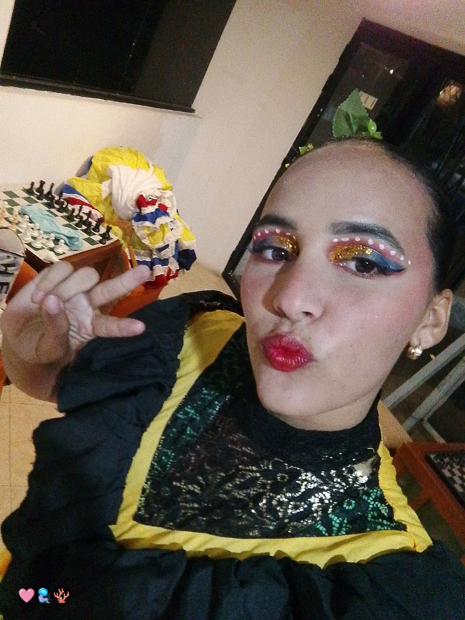 An indoor, eye-level, slightly high-angle photograph depicts a young woman posing for a selfie in Playa El Agua, Venezuela. The woman, with dark hair pulled back, is making a "kiss" face with her vibrant red-lipsticked mouth and forming a peace sign with her raised left hand. Her eyes feature elaborate theatrical makeup, including gold glitter, white dots, and blue and pink eyeshadow. She wears a black top with sheer, patterned lace fabric over a bright yellow garment, and black gathered sleeves. A gold hoop earring is visible in her right ear. In the background to her left, a wooden chessboard with black and white chess pieces is visible on a surface. Behind the chessboard, a pile of colorful fabrics, including yellow, white, blue, and red components, is stacked. The upper background consists of a dark, rectangular area, possibly a window or shelf, against a light-colored wall. The lower-left foreground displays three digital emojis: a pink heart, a blue jellyfish, and a red coral.