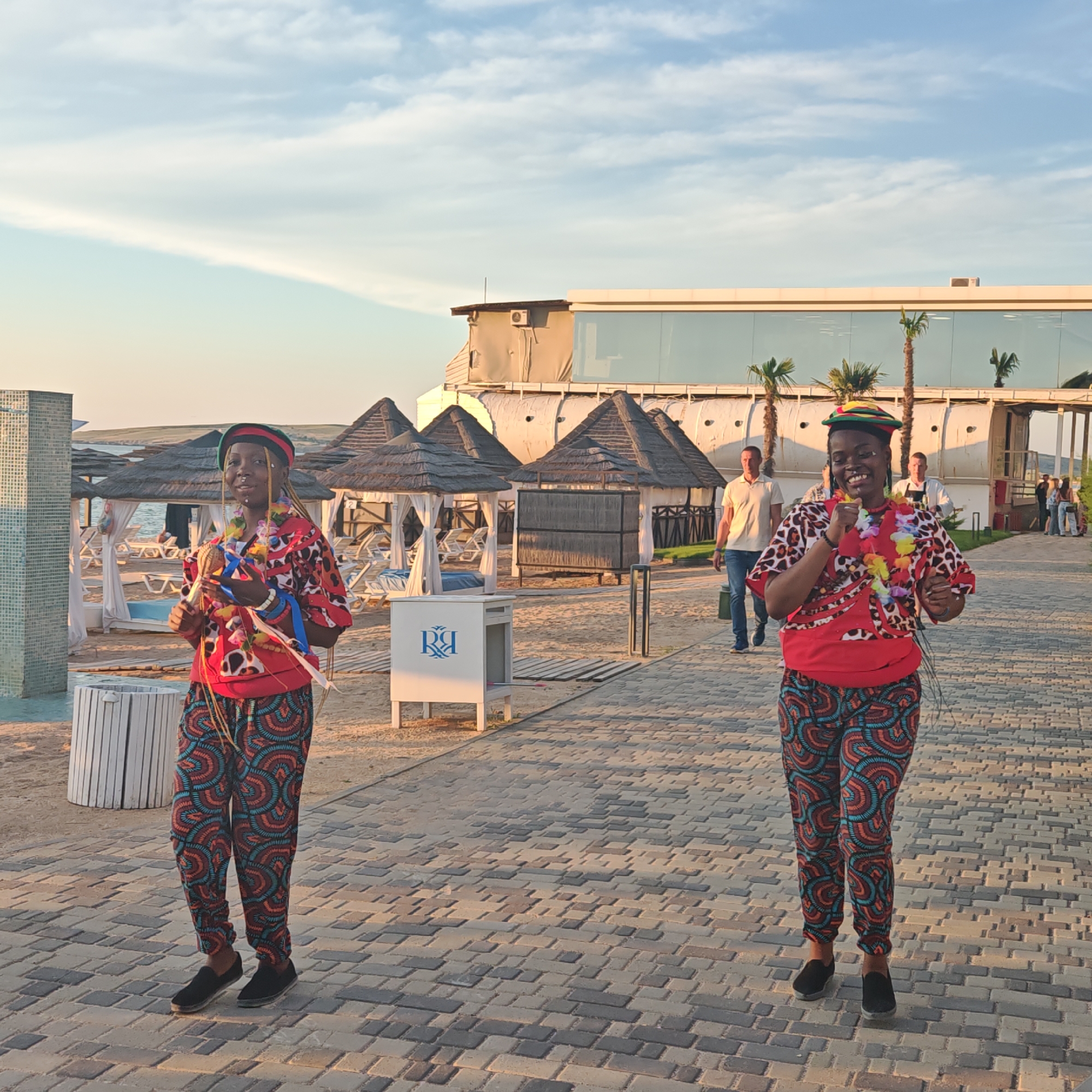 The image shows two women dressed in traditional attire and leis, appearing to be performers or dancers, walking on a paved walkway near a beach. In the background, there are thatched-roof cabanas, lounge chairs, and a modern building with large glass windows reflecting the sky. The environment suggests a resort or entertainment venue on a sunny day, possibly during a performance. The city of Мысовское сельское поселение, Ukraine is a rural settlement in the Odesa Oblast of Ukraine.