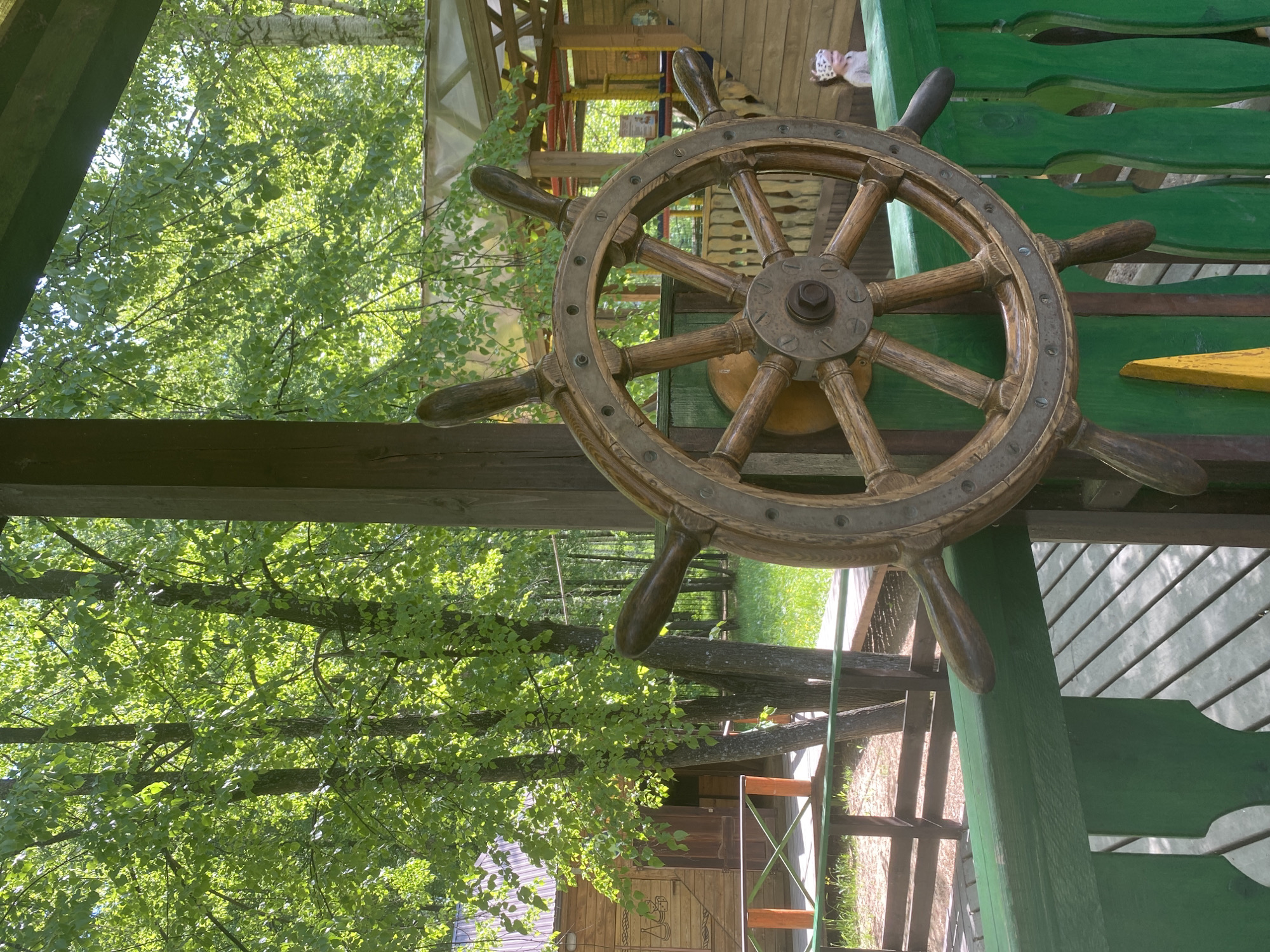 The image shows a wooden ship's wheel mounted on a green wooden structure in an outdoor setting. The wheel has a brown wooden frame with metal accents and eight handles. The backdrop consists of lush green trees and foliage, suggesting a park or garden. The scene appears to be outdoors, with the wooden structure potentially part of a playground or decorative element. Given the prompt, this is likely located in Chaikovsky, Russia.
