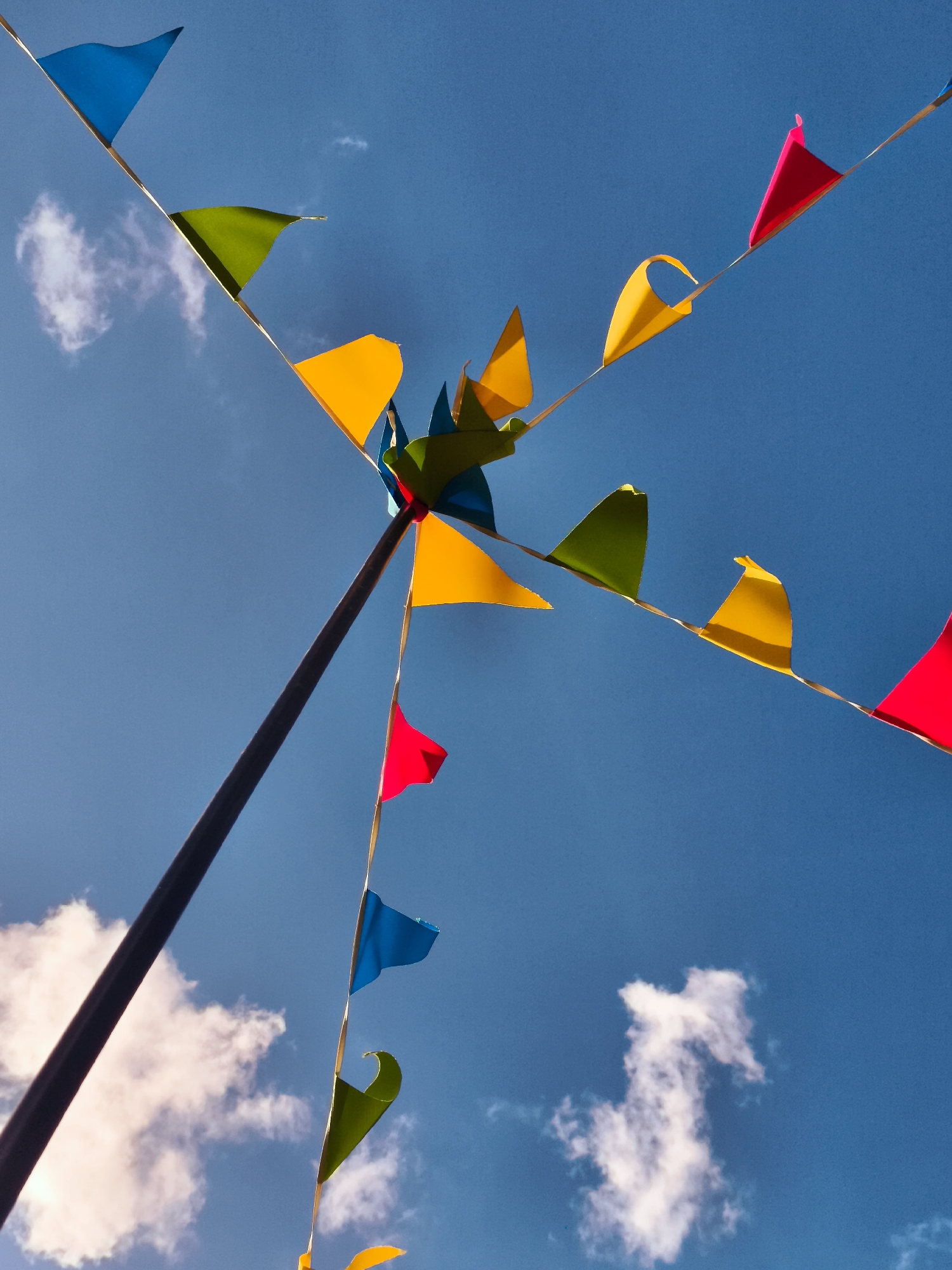 The image is a low-angle shot of colorful pennant flags strung across a bright blue sky with scattered white clouds. A dark pole extends diagonally from the bottom left corner upwards, supporting a cluster of pennants that fan out in various directions. The pennants are triangular and in hues of blue, green, yellow, and red, with some appearing slightly creased or curved by the wind. The scene suggests an outdoor event or celebration. No people are visible. The context of Gdańsk, Poland, is not visually evident in the image itself.