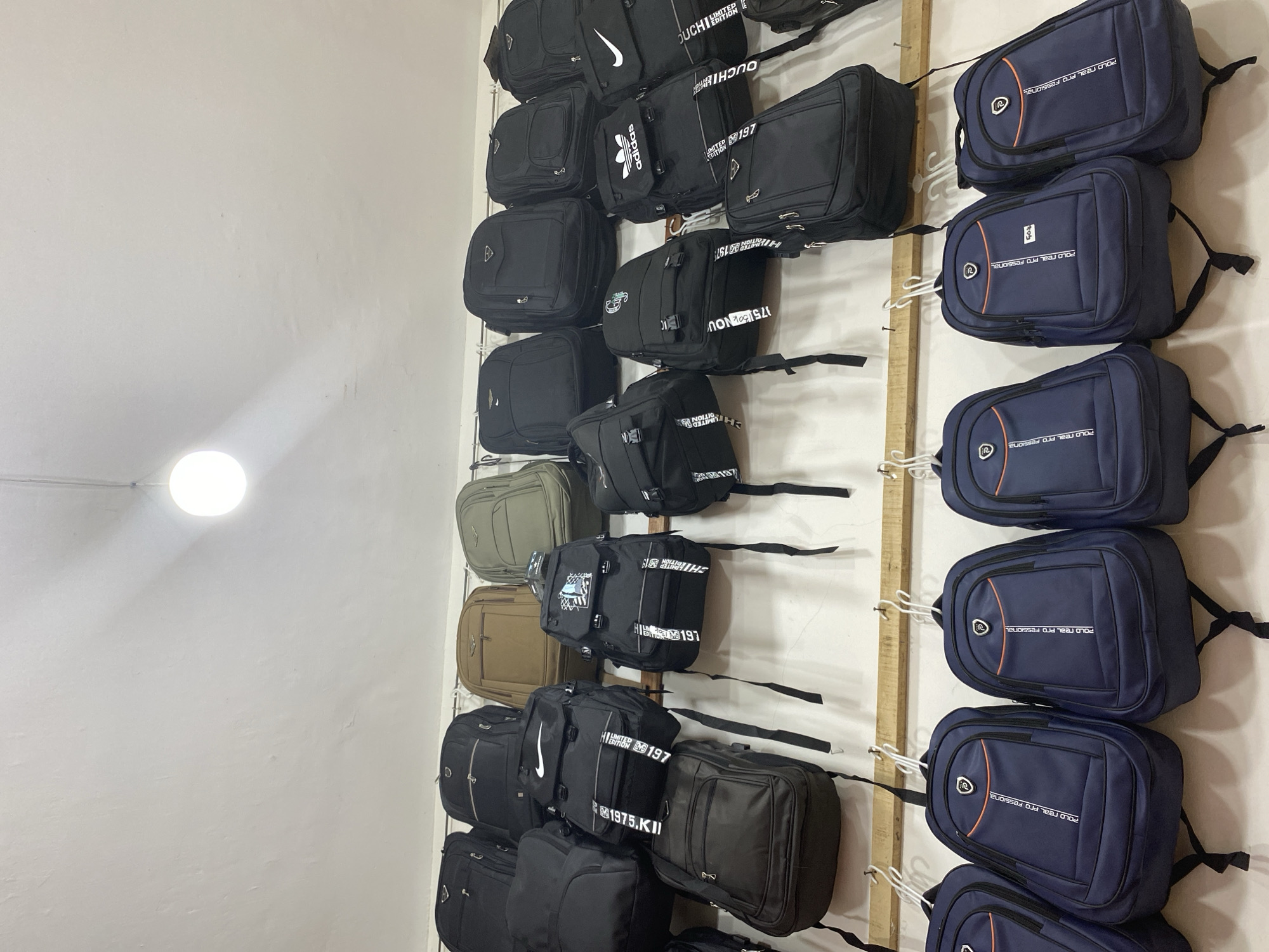 This image captures a retail display of backpacks in what appears to be a market stall in Lamteumen Barat, Indonesia. The backpacks are predominantly black, with a few in navy blue, tan, and olive green. They are arranged in rows, hung on hooks and shelves against a white wall. Some backpacks have visible branding, including Nike, Adidas, and what looks like a "Polo Real Pro Fessional" logo. One black backpack in the center has "LIMITED EDITION 1975 KII" printed on a strap. Below the higher rows of backpacks, there are several navy blue backpacks with orange accents and the "Polo Real Pro Fessional" branding. The lighting suggests an indoor environment, possibly a shop or market. There are no people visible in the image, only the merchandise.
